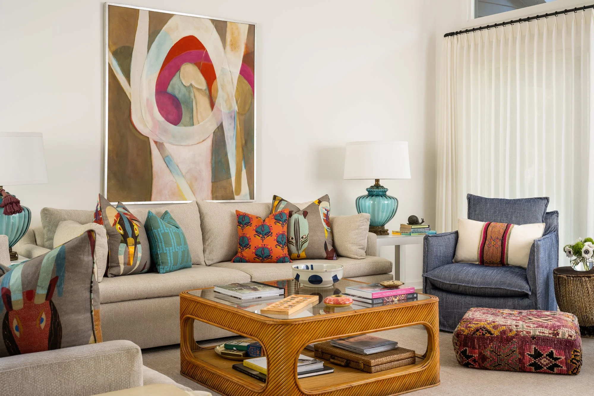 K. Interiors Design Work - Living room with beige sofa and blue armchair, colorful pillows, wooden coffee table, large abstract art on white wall, table lamps, and window with cream curtains.