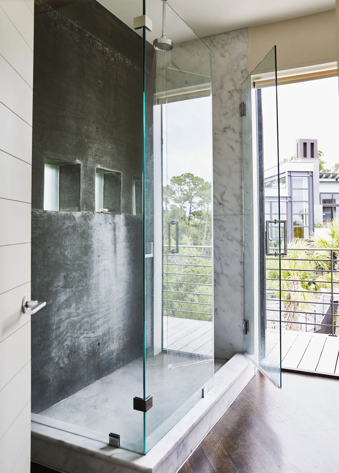 K. Interiors Design Work - Modern glass shower enclosure with a rain shower head, marble and textured wall, and open balcony with greenery outside.