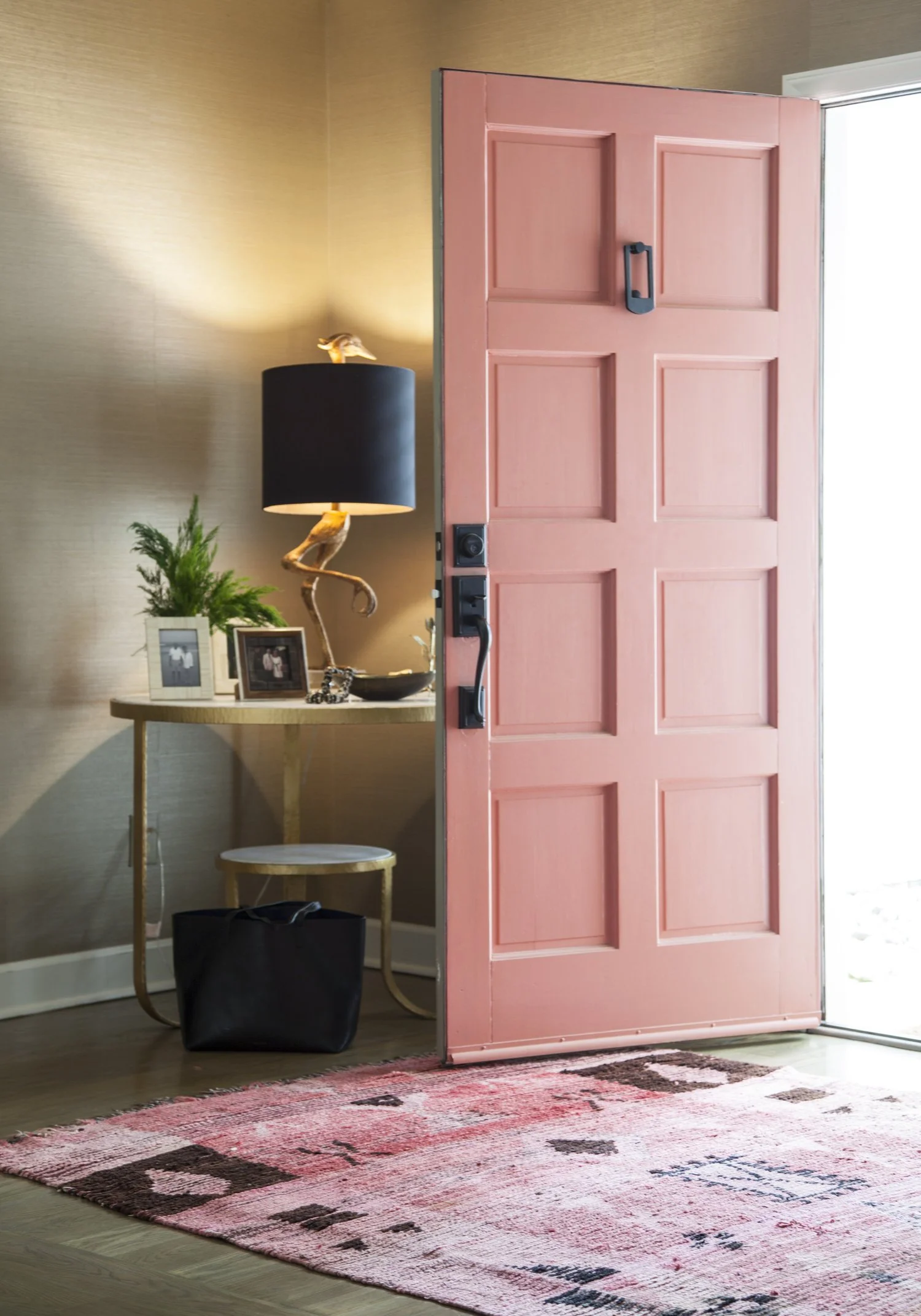 K. Interiors Design Work - Open pink front door revealing a cozy entryway with a wooden side table, black lampshade with a seagull figurine, picture frames, green plant, black bag, and a red and pink patterned rug on the hardwood floor.