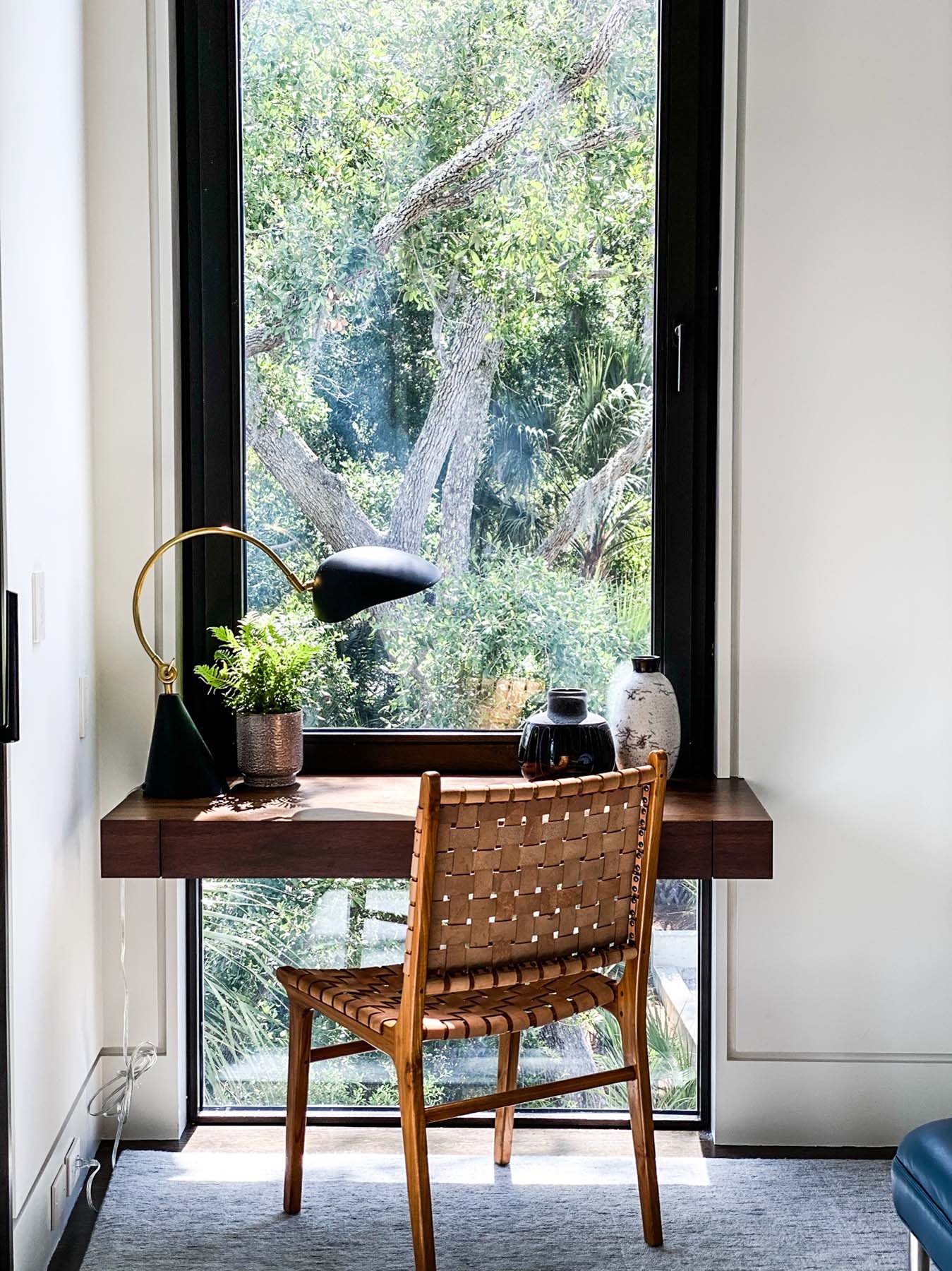 K. Interiors Design Work - A wooden desk with a black adjustable desk lamp, potted fern, and decorative vases, positioned in front of a large window overlooking green trees outside.