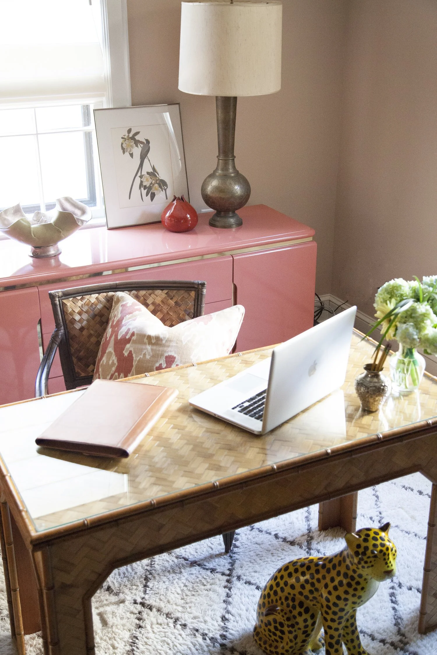 K. Interiors Design Work - A home office with a wooden desk, a pink sideboard, a table lamp, a framed bird and flower art, a small red vase, a laptop, a notebook, a flower vase, and a leopard sculpture on a rug.
