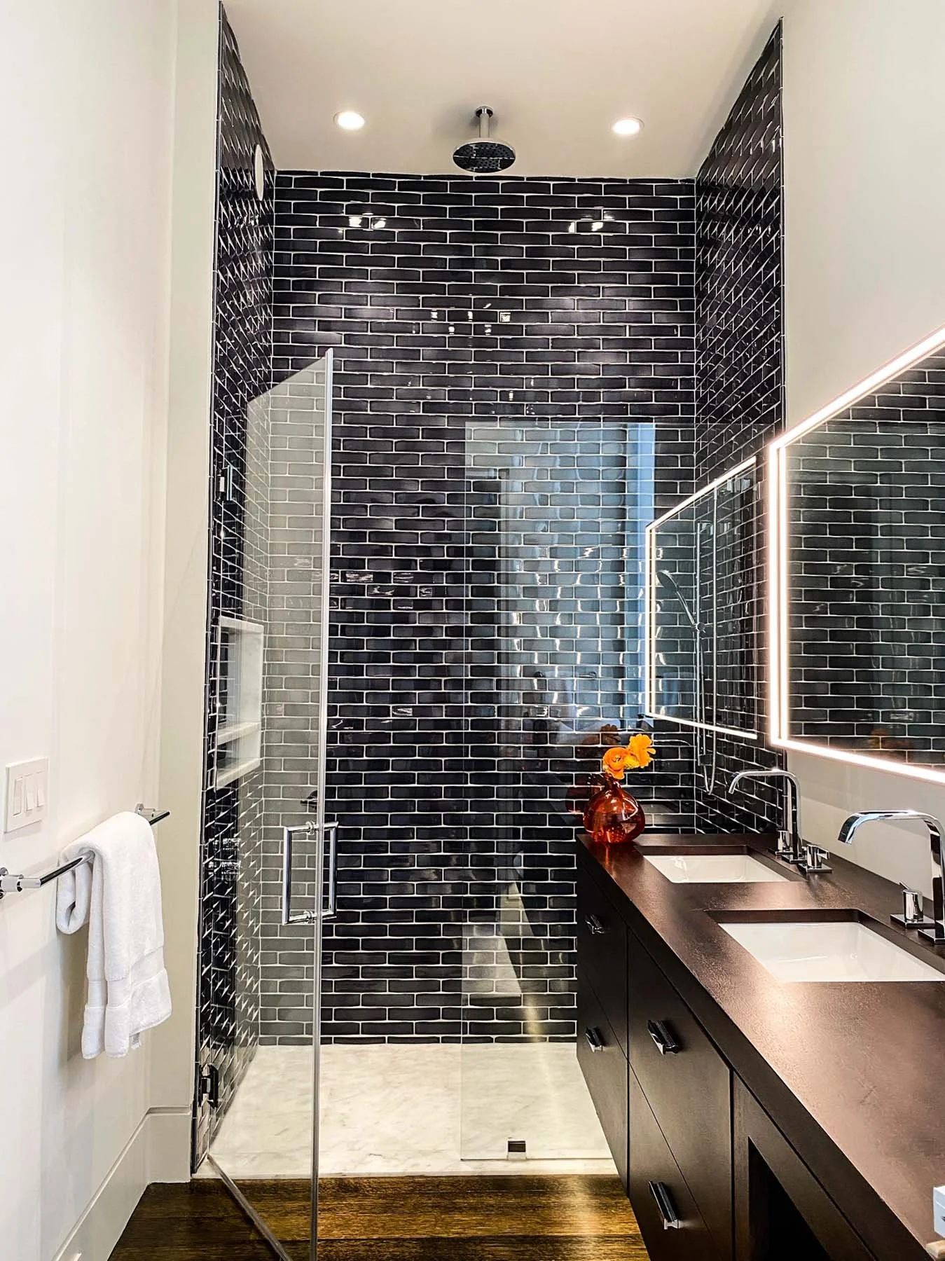 K. Interiors Design Work - Modern bathroom with black brick-tile shower, white walls, wooden floor, black vanity with twin sinks, backlit mirror, decorative orange flowers in a brown vase.