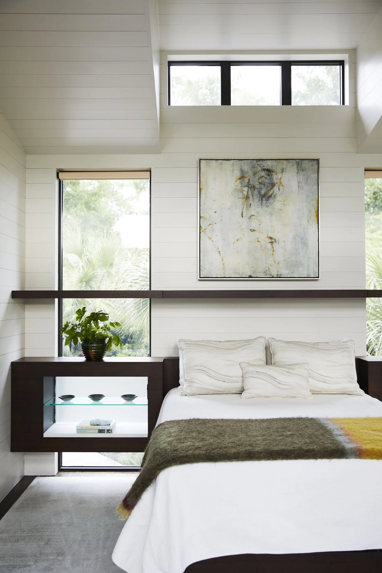 K. Interiors Design Work - A modern bedroom with white walls, large windows, a bed with white bedding, pillows, and a brown throw, a potted plant on a side table, and an abstract wall art piece.