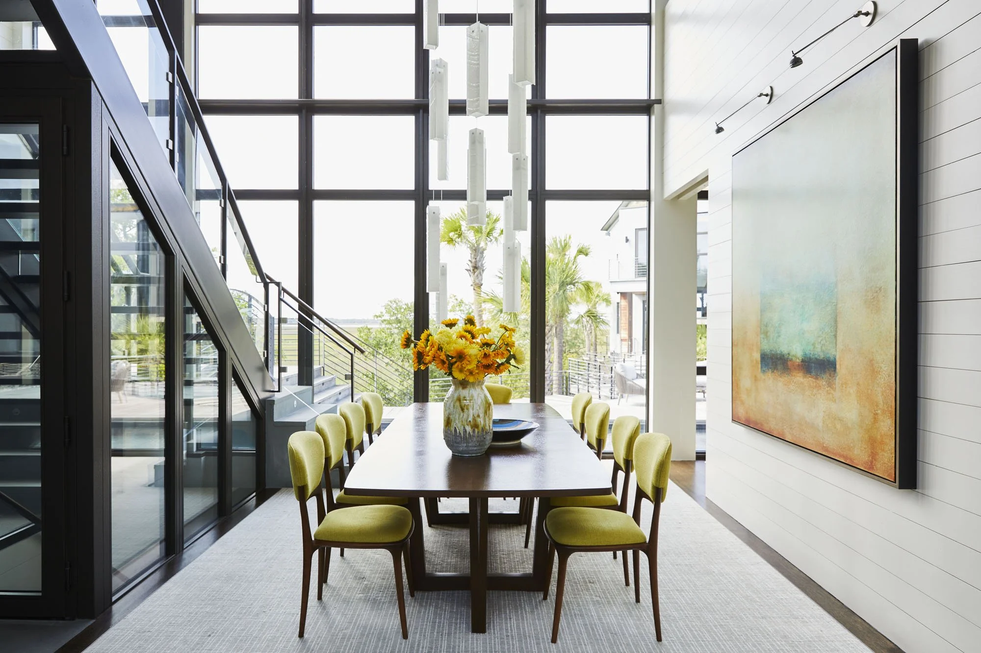K. Interiors Design Work - Modern dining room with a large wooden table, ten green cushioned chairs, a flower arrangement on the table, a large abstract painting on the wall, and floor-to-ceiling windows letting in natural light.
