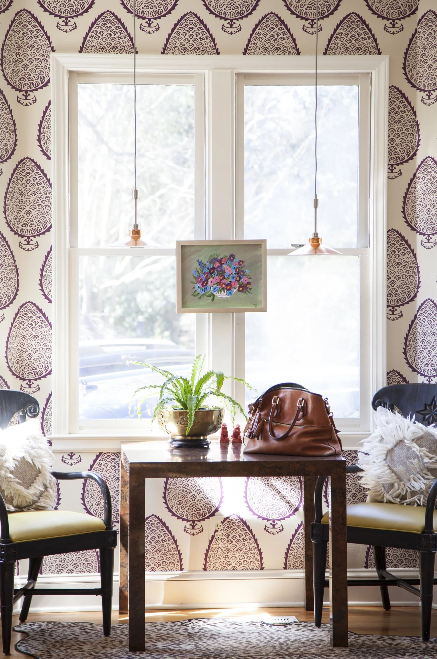 K. Interiors Design Work  - Interior of a room with a large window, wooden table with a potted fern. Two patterned chairs with fringed pillows. A painting of a colorful floral bouquet hangs on the window, with patterned wallpaper and pendant lights.