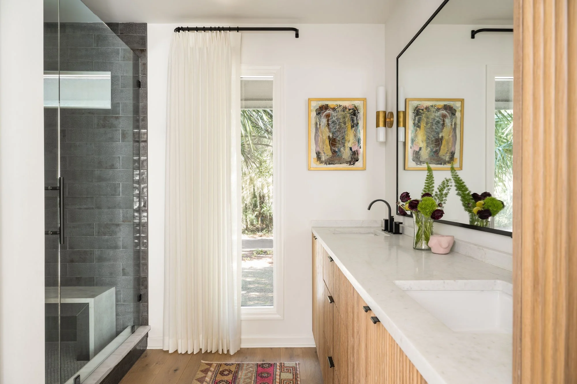K. Interiors Design Work - Modern bathroom with white walls, a marble countertop with a built-in sink, and wooden cabinetry. There is a black faucet, artwork on the wall, a window with a white curtain, and a walk-in shower with dark gray tiles.
