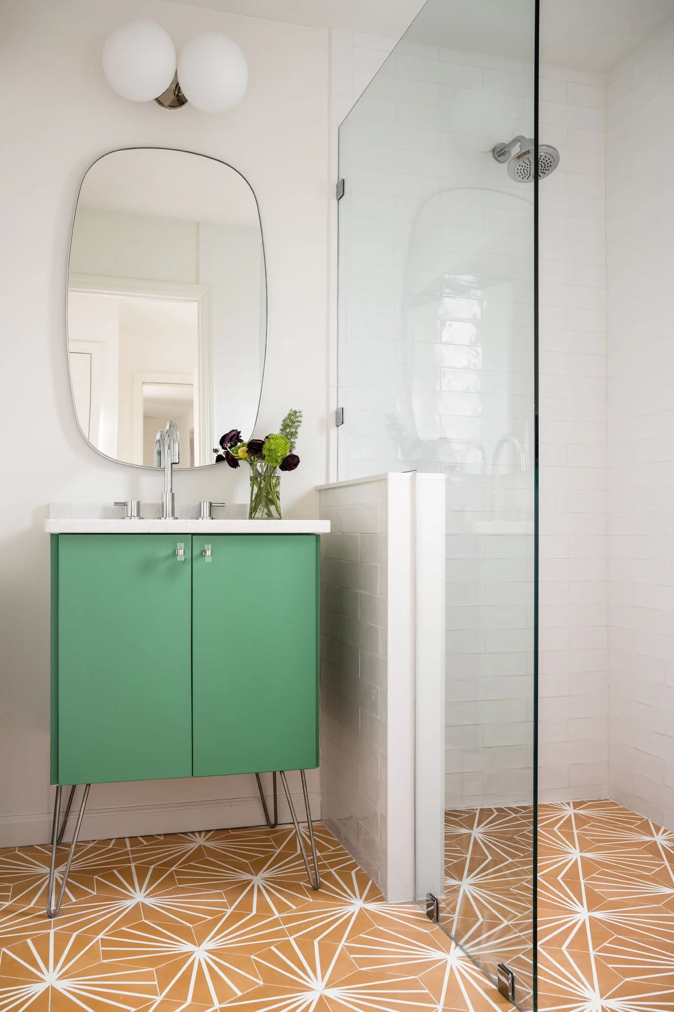 K. Interiors Design Work - Modern bathroom with a mint green vanity, white countertop, oval mirror, glass shower door, white tiled shower, and patterned tile flooring.