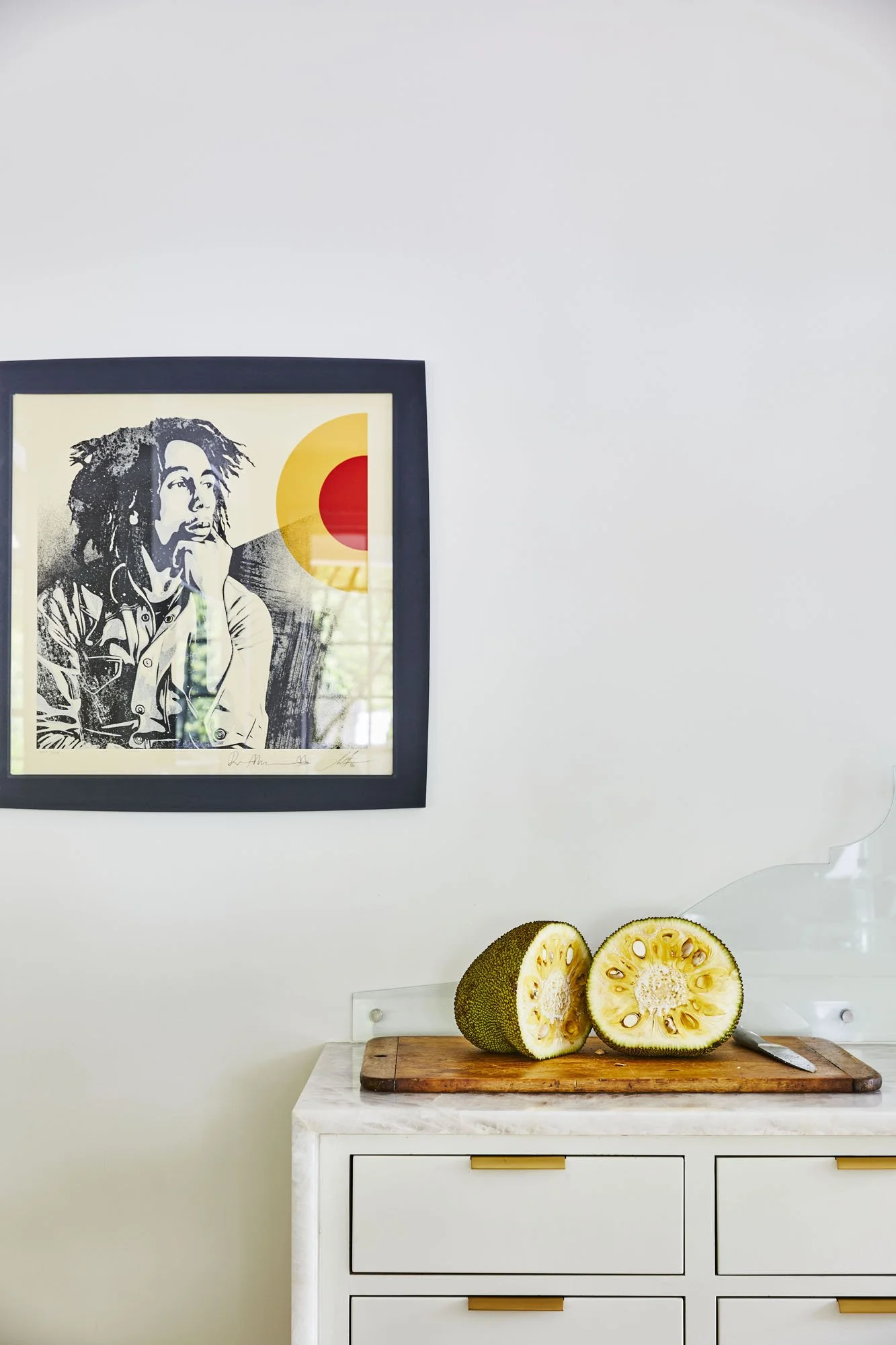 K. Interiors Design Work - A white cabinet with a wooden cutting board on top, holding a sliced kiwano melon and a knife. A framed black-and-white portrait of Bob Marley hangs on the white wall above.