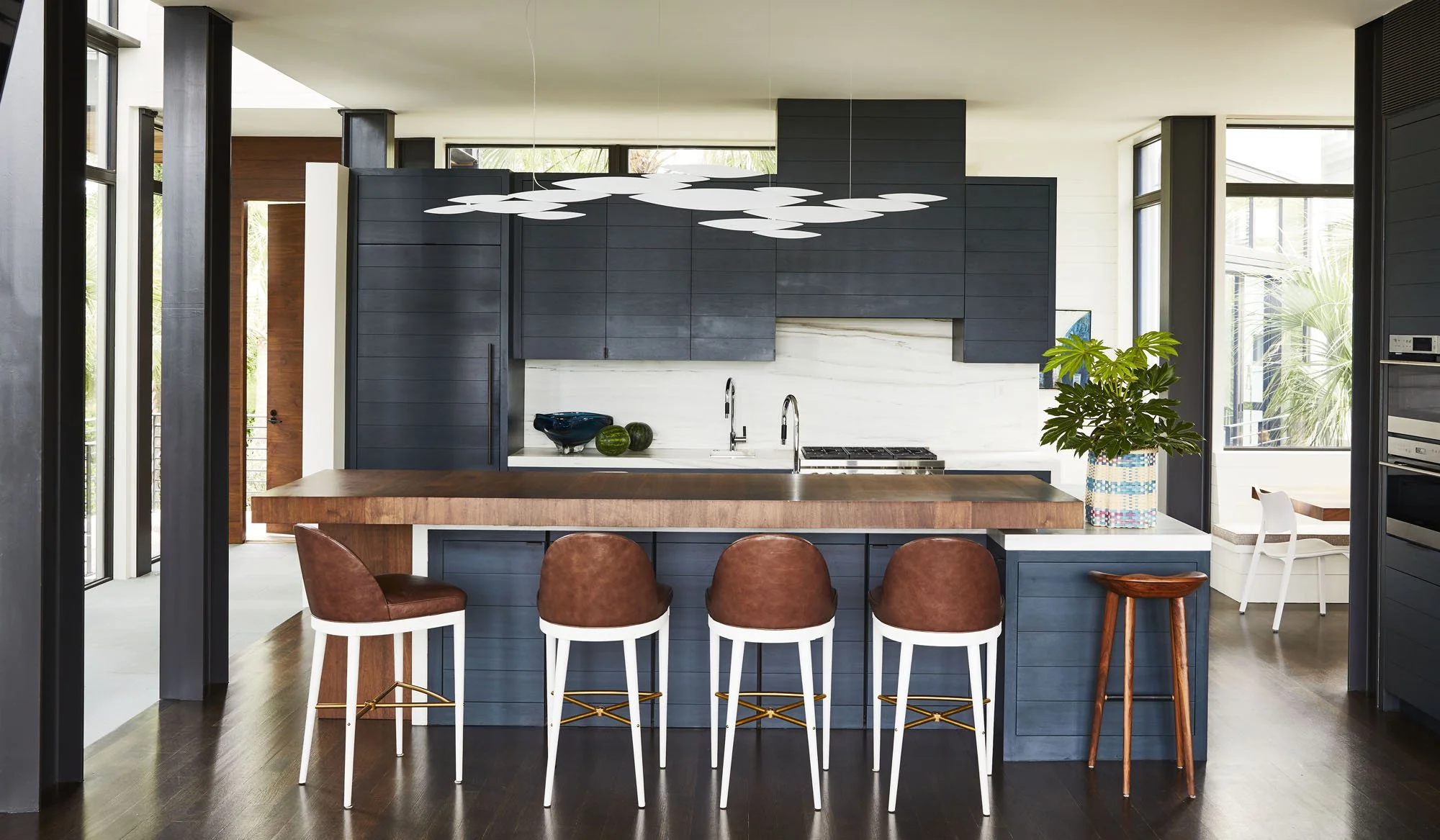 K. Interiors Design Work - Modern kitchen with navy blue cabinets, a wooden island, four brown and white barstools, and a large potted plant on the counter, with bright windows and contemporary lighting.