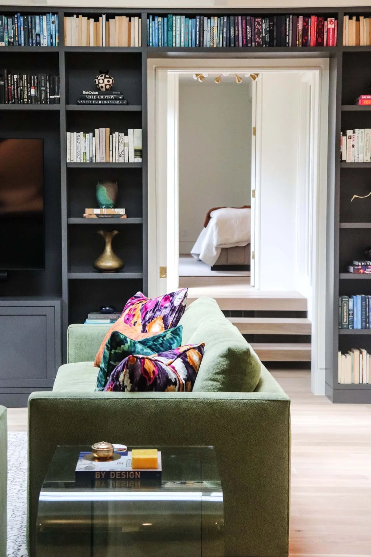 K. Interiors Design Work - A living room with a green sofa decorated with colorful pillows, a glass-top coffee table with a book and decorative items, and a built-in bookshelf filled with books and decorative objects. 