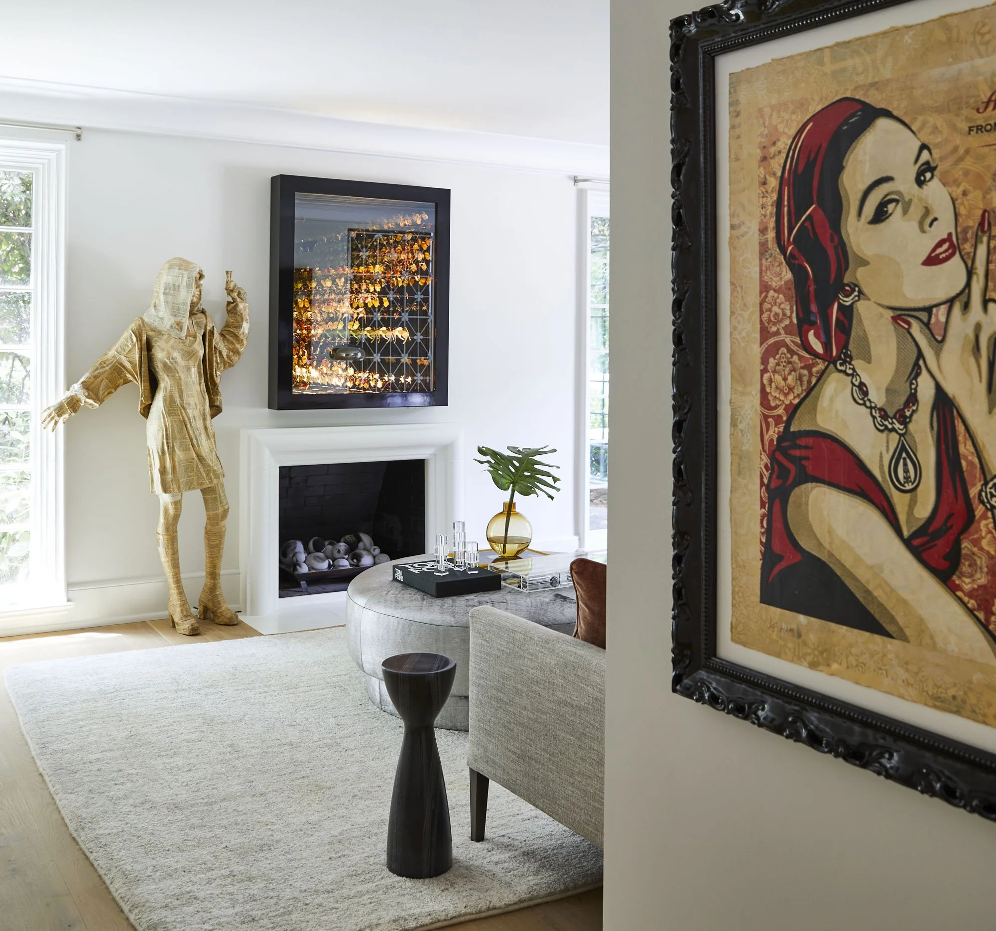 K. Interiors Design Work - Modern living room with beige sofa, decorative vase with leaf, artwork of woman with red hair, sculpture of a figure in gold, white fireplace, and fire in a black-framed glass cabinet.