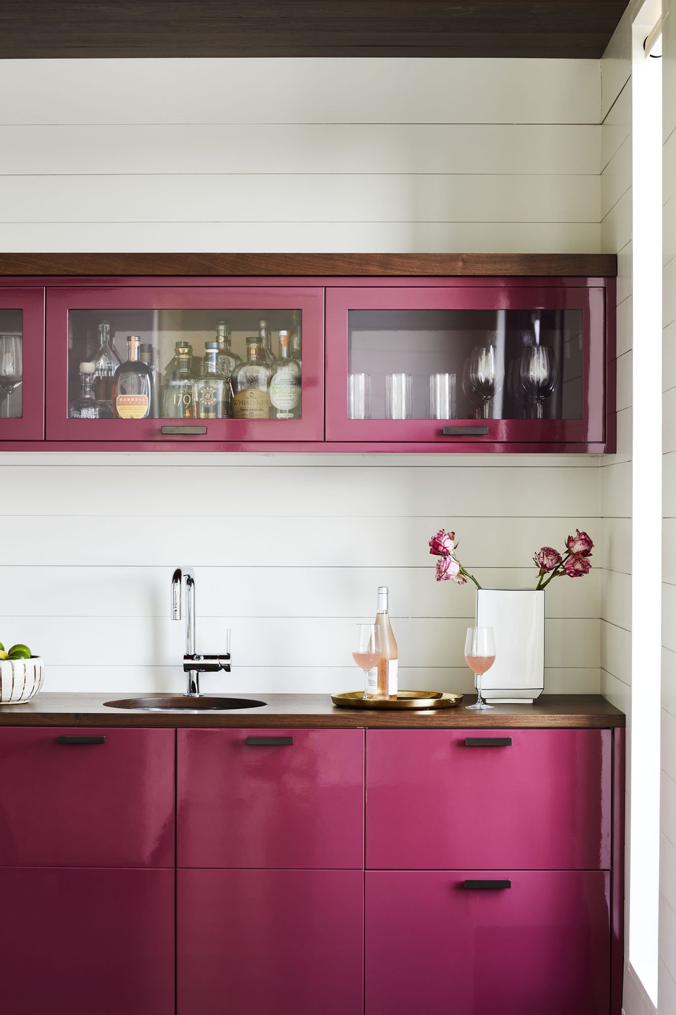 K. Interiors Design Work - Pink cabinets in a kitchen with a white shiplap wall, a small sink, a bottle of rosé, two glasses of rosé, a white vase with pink flowers, and a gold tray.