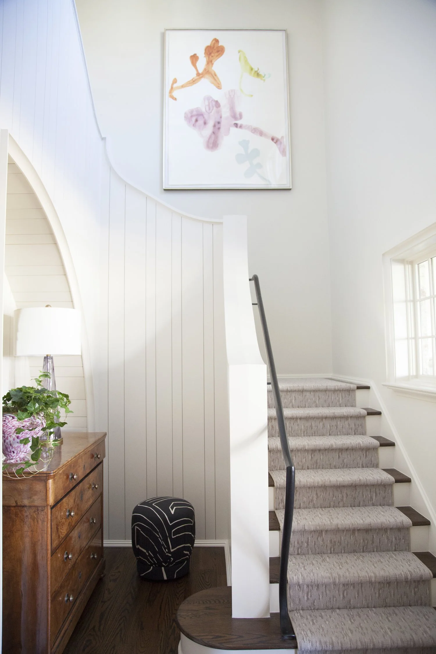 K. Interiors Design Work - Interior staircase with beige carpeting, white walls, a window, a wooden console table with a lamp and plants, and a large abstract watercolor painting above.