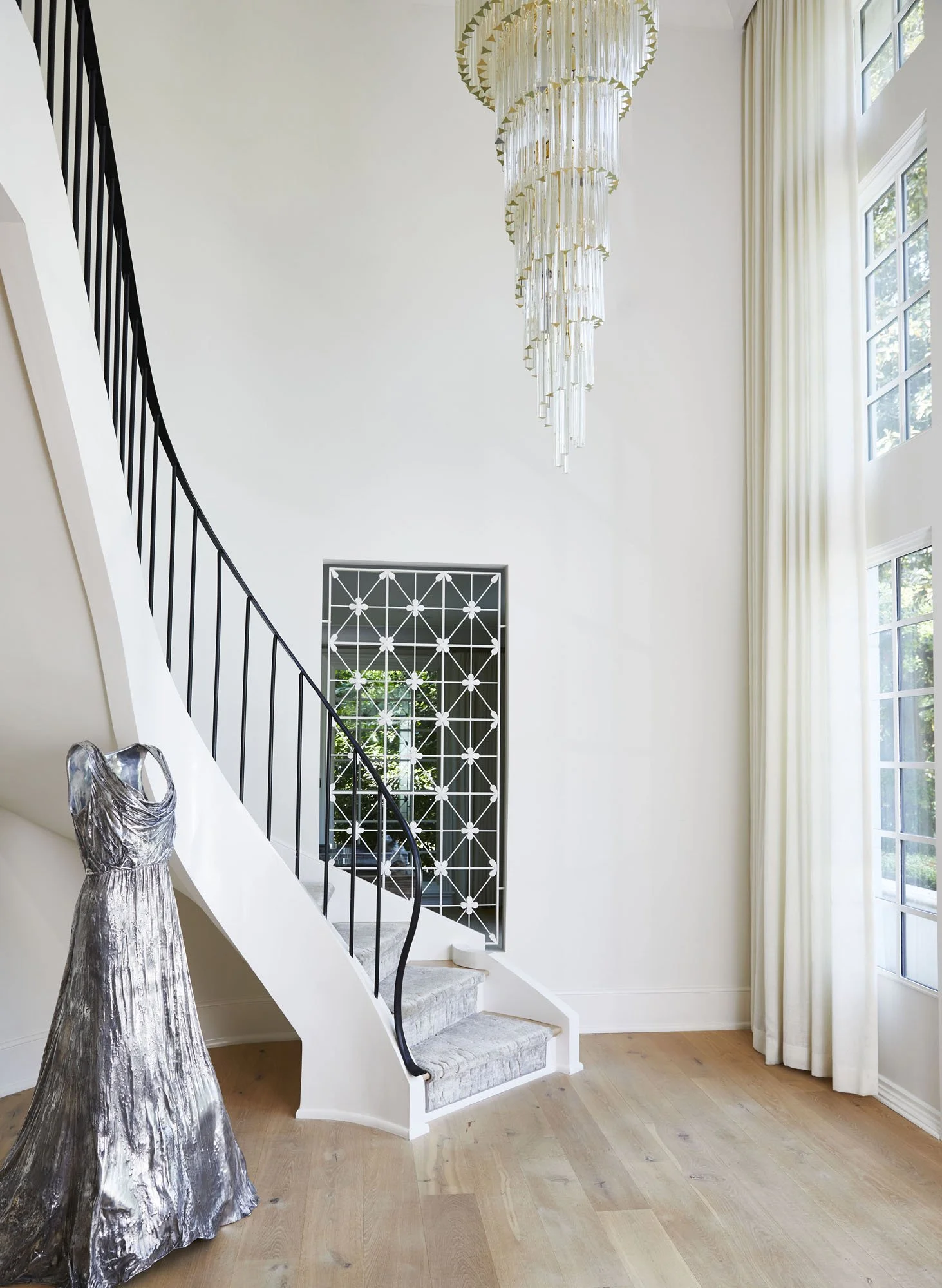 K. Interiors Design Work - Interior of a modern house with a staircase, a chandelier hanging from the ceiling, large windows with curtains, a decorative artwork on the wall, and a metallic sculpture of a dress.