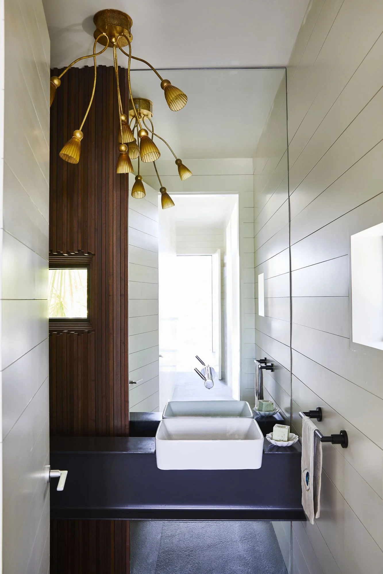 K. Interiors Design Work - Modern bathroom with a white rectangular vessel sink, a black countertop, a large mirror, a gold chandelier, and wooden paneling on the wall.