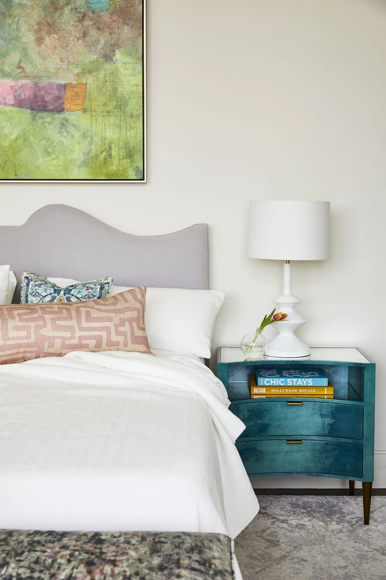 K. Interiors Design Work - A bedroom featuring a bed with white linens, decorative pillows, and a light gray headboard. Beside the bed is a teal nightstand with a glass vase holding a flower, a white table lamp, and books.
