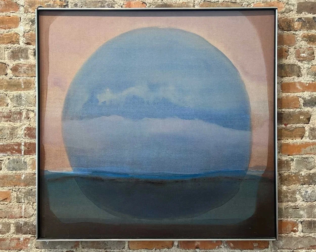 Abstract artwork with a large blue circle over a landscape of clouds and water, framed and mounted on a brick wall.