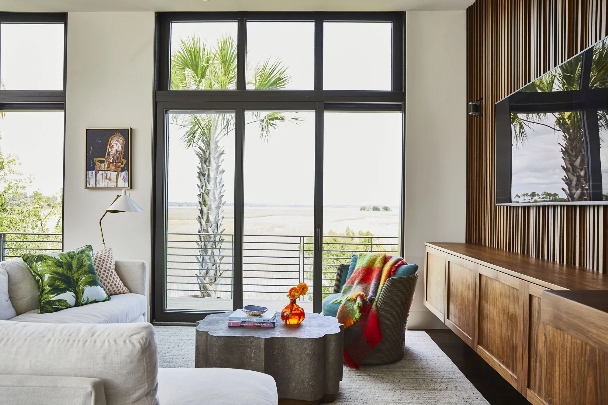 K. Interiors Design Work - Living room with large windows, palm trees outside, a white sofa with green leaf pillows, a modern floor lamp, a concrete coffee table with books and an orange vase, a colorful blanket on a chair, and wooden sideboard.