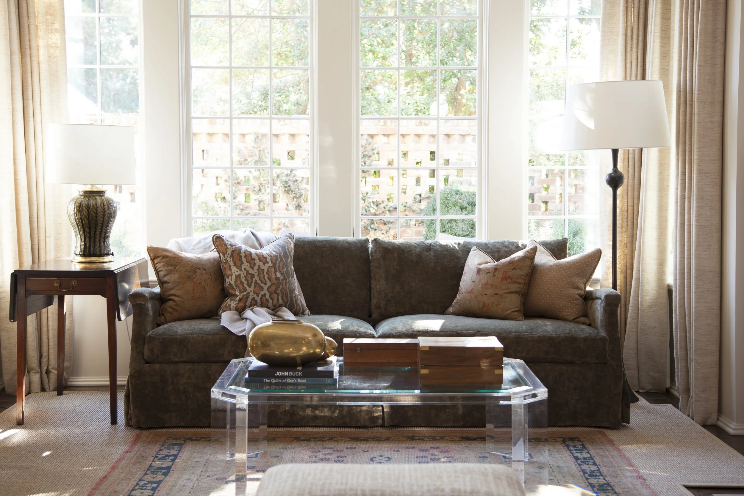 K. Interiors Design Work - Living room with a gray sofa, decorative pillows, a glass coffee table with books and a gold decorative object, a side table with a lamp, and large windows with curtains that let in natural light.