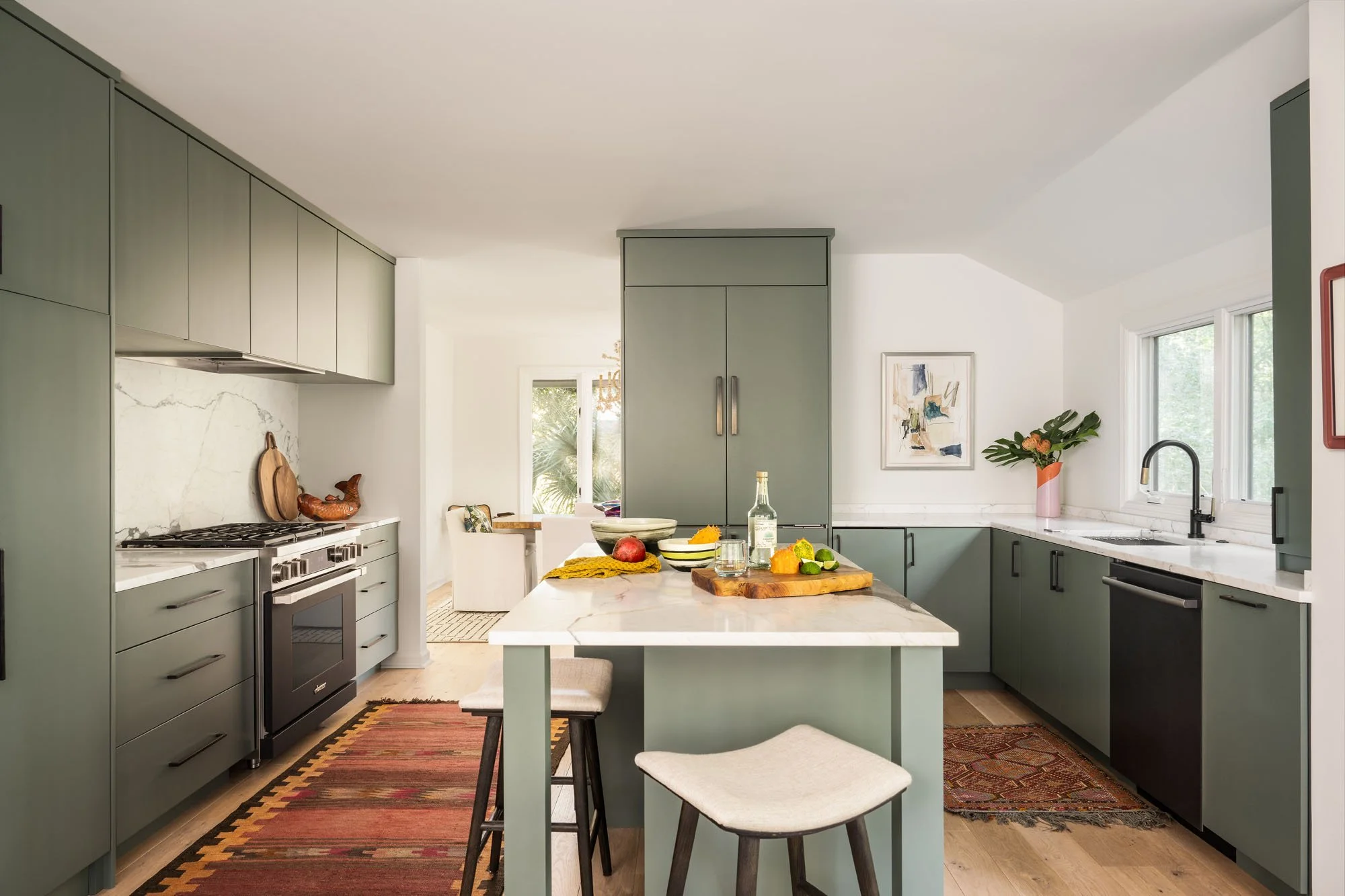 K. Interiors Design Work - Modern kitchen with green cabinetry, white marble countertops, and a central island with stools; decorated with artwork, flowers, and food items.