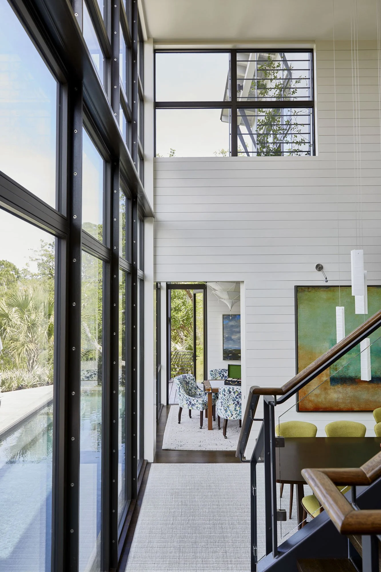 K. Interiors Design Work - Interior view of a modern house with large glass windows, overlooking a backyard with a pool, featuring a staircase, colorful artwork, and a dining area with patterned chairs.
