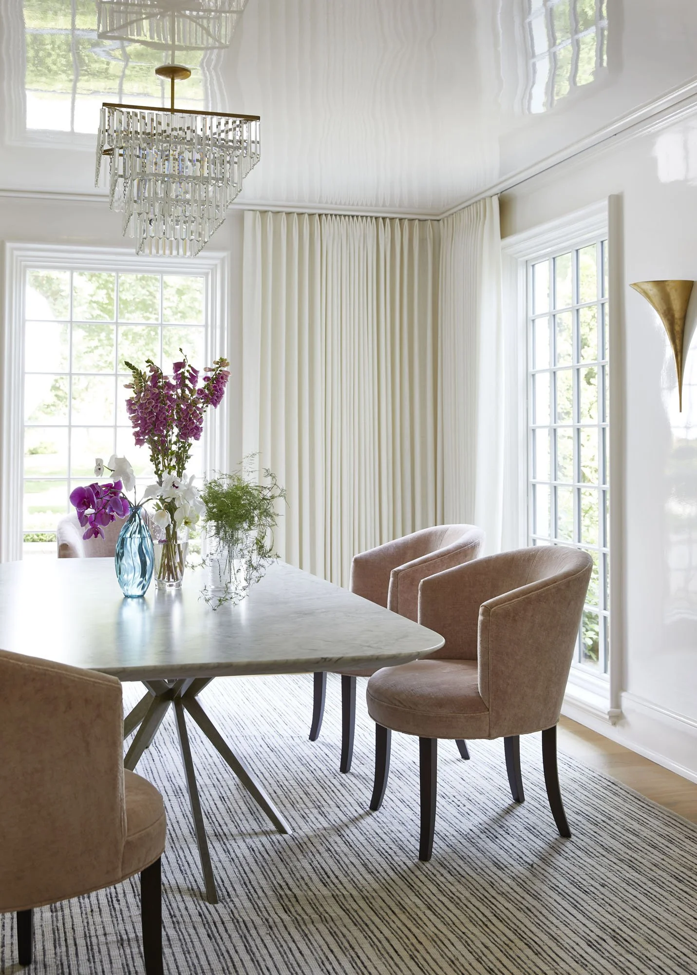 K. Interiors Design Work - A bright dining room with large windows, beige chairs around a white marble table, and a chandelier hanging from the ceiling. There are vases with flowers on the table and long white curtains on the windows.