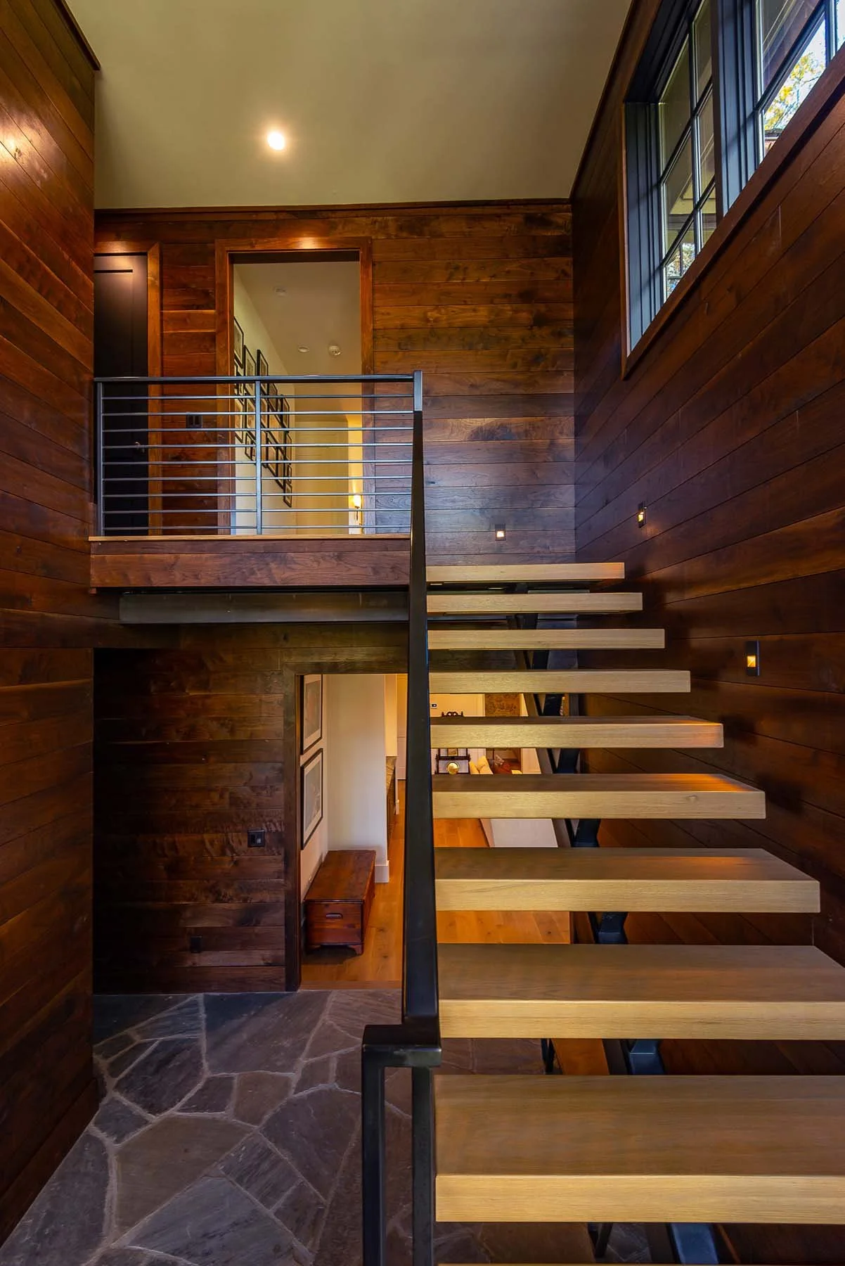 K. Interiors Design Work - Interior view of a modern wooden staircase with open risers and black metal railing in a home with dark wood walls, natural light from large windows, and stone flooring.