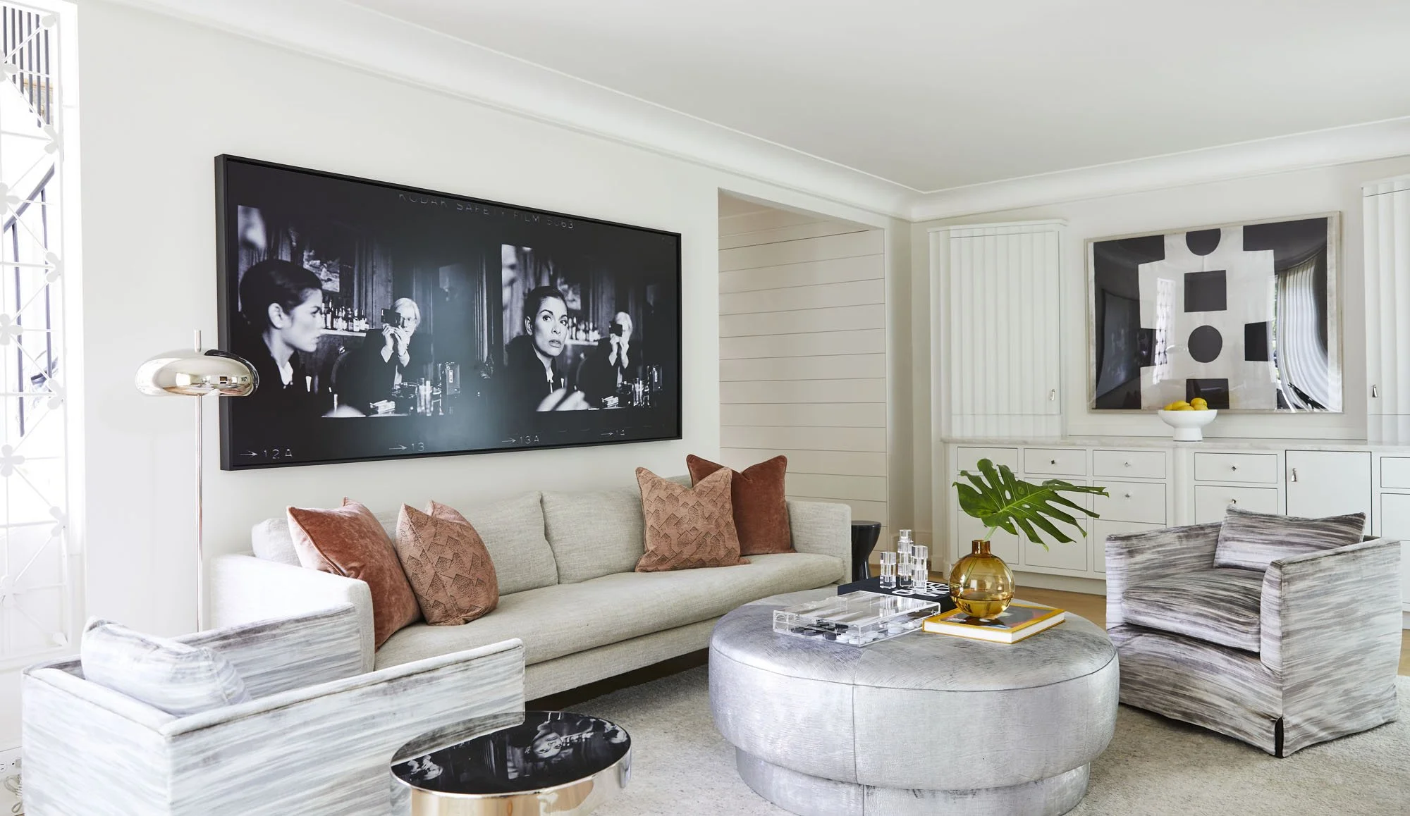 K. Interiors Design Work - Living room with beige sofa with pink pillows, matching armchair, round silver ottoman with a glass tray, yellow vase with a large green leaf, white sideboard with a modern black and white artwork, and white walls.