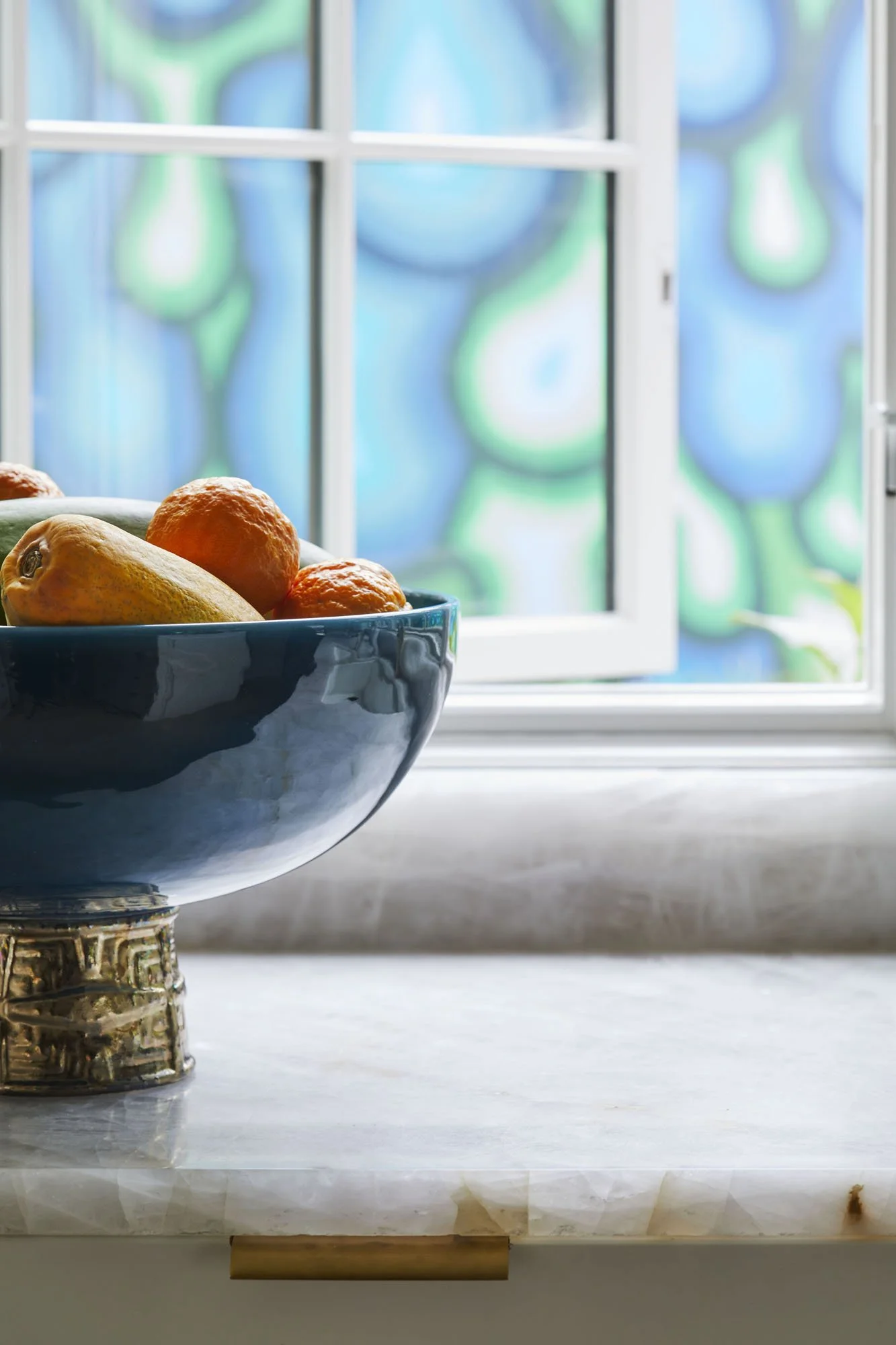 K. Interiors Design Work - A blue bowl filled with oranges, tangerines, and a yellow pear sitting on a marble countertop next to a window with colorful abstract stained glass art outside.