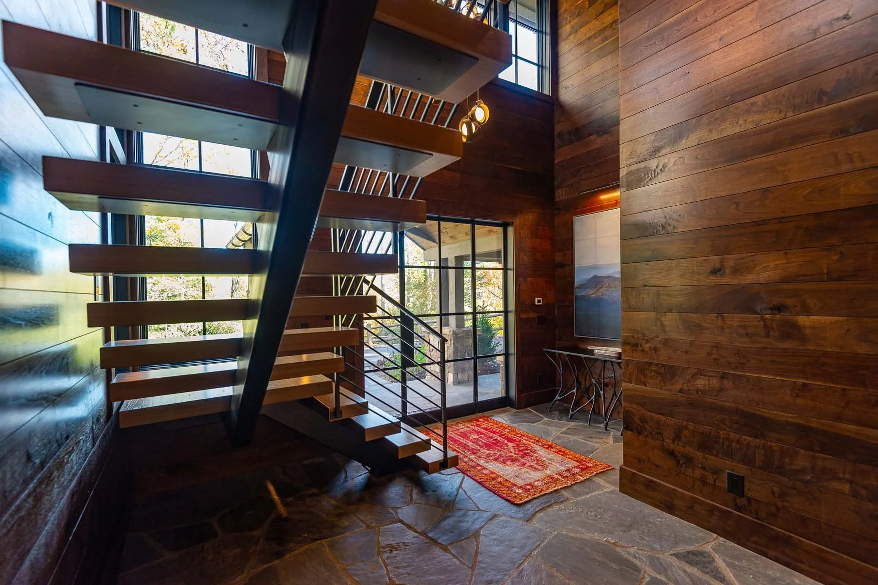 K. Interiors Design Work - Interior view of a modern home staircase with wooden steps and metal railing, large glass windows showing trees outside, a small decorative table, a painting, and a colorful rug on a stone floor.