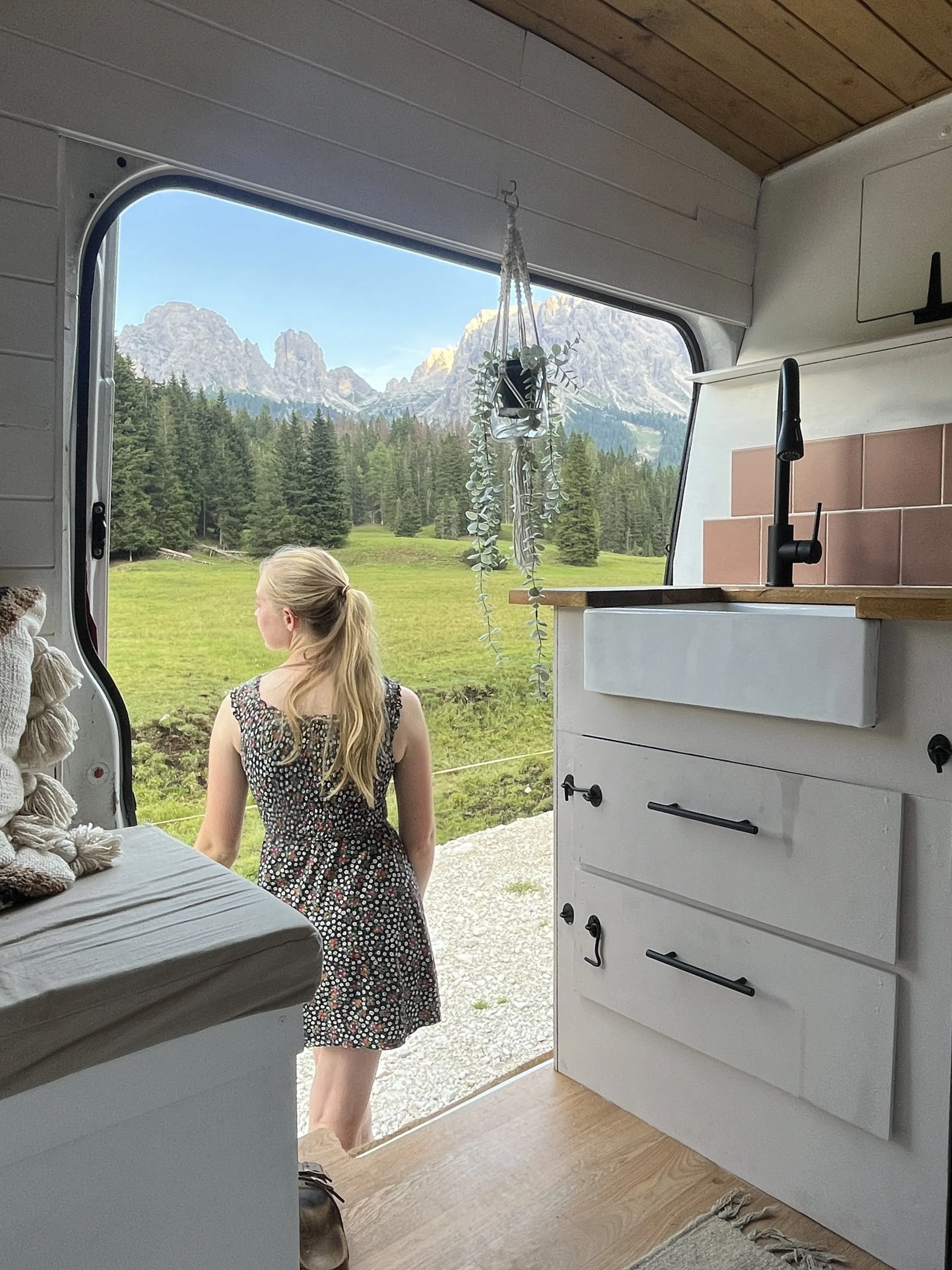 Taken from inside a converted van looking out through the open sliding door. A blonde woman with her hair in a ponytail stands at the open door looking away from the camera towards a view of pine trees and rocky mountain scenery in the Dolomites.