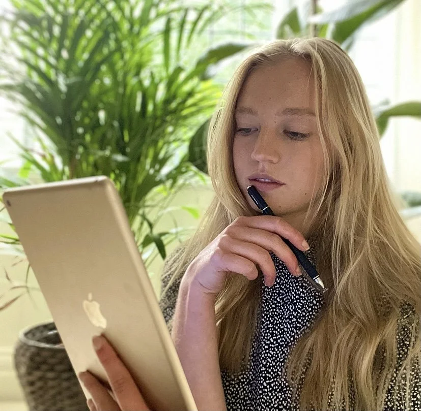 A blonde woman holds an iPad and stylus, holding the stylus up to her face thoughtfully. Behind her is an array of houseplants.