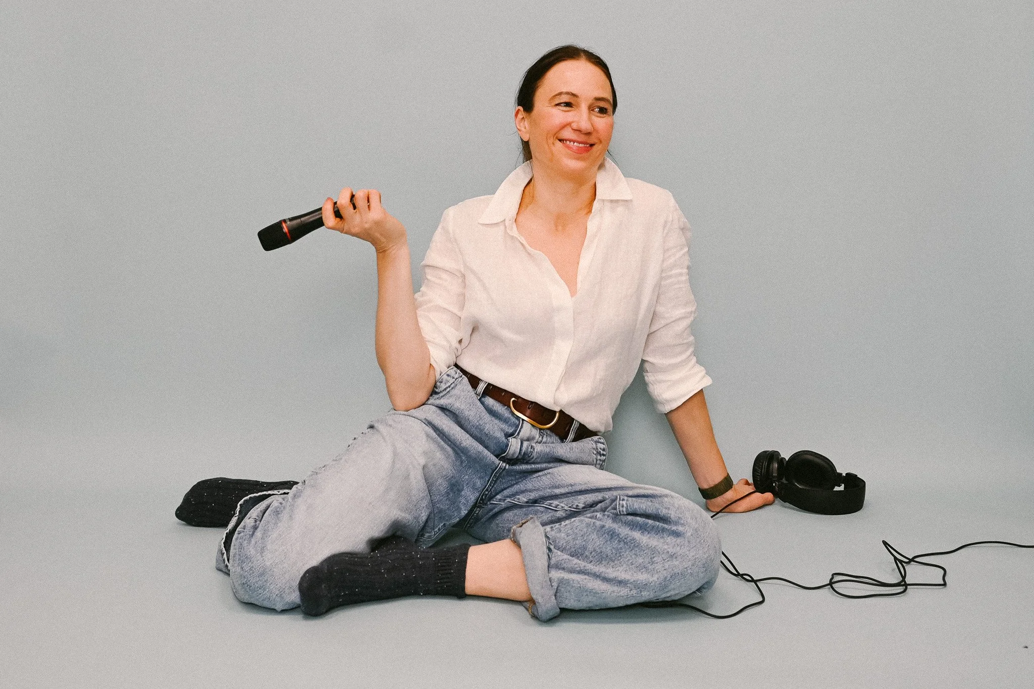 A woman sitting on the floor with a vintage microphone in her hand, smiling, with headphones on the ground beside her, against a plain gray background.