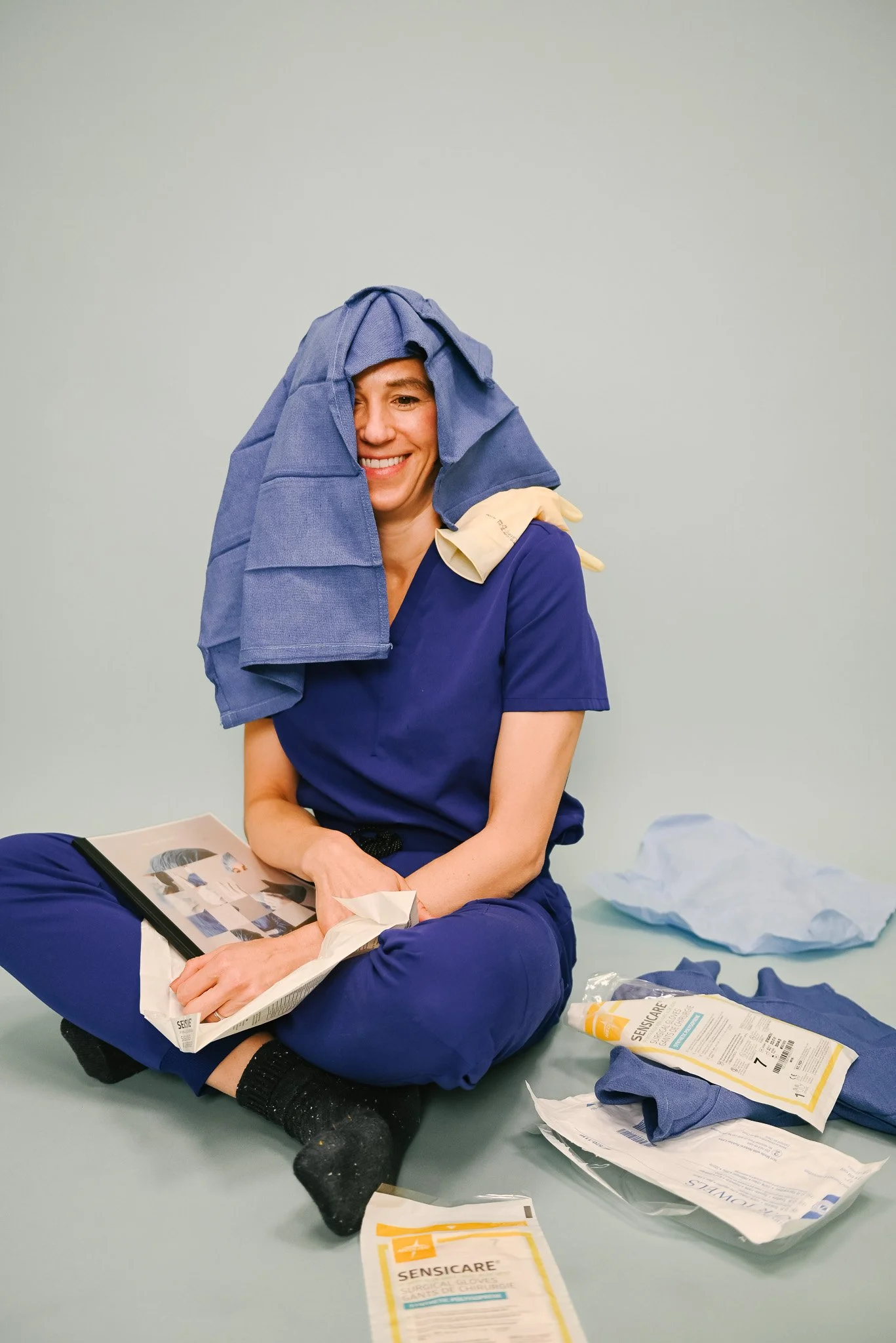 A woman sitting on the floor, smiling with medical supplies around her, wearing blue scrubs and a blue cloth covering her head, with a yellow glove on her shoulder.