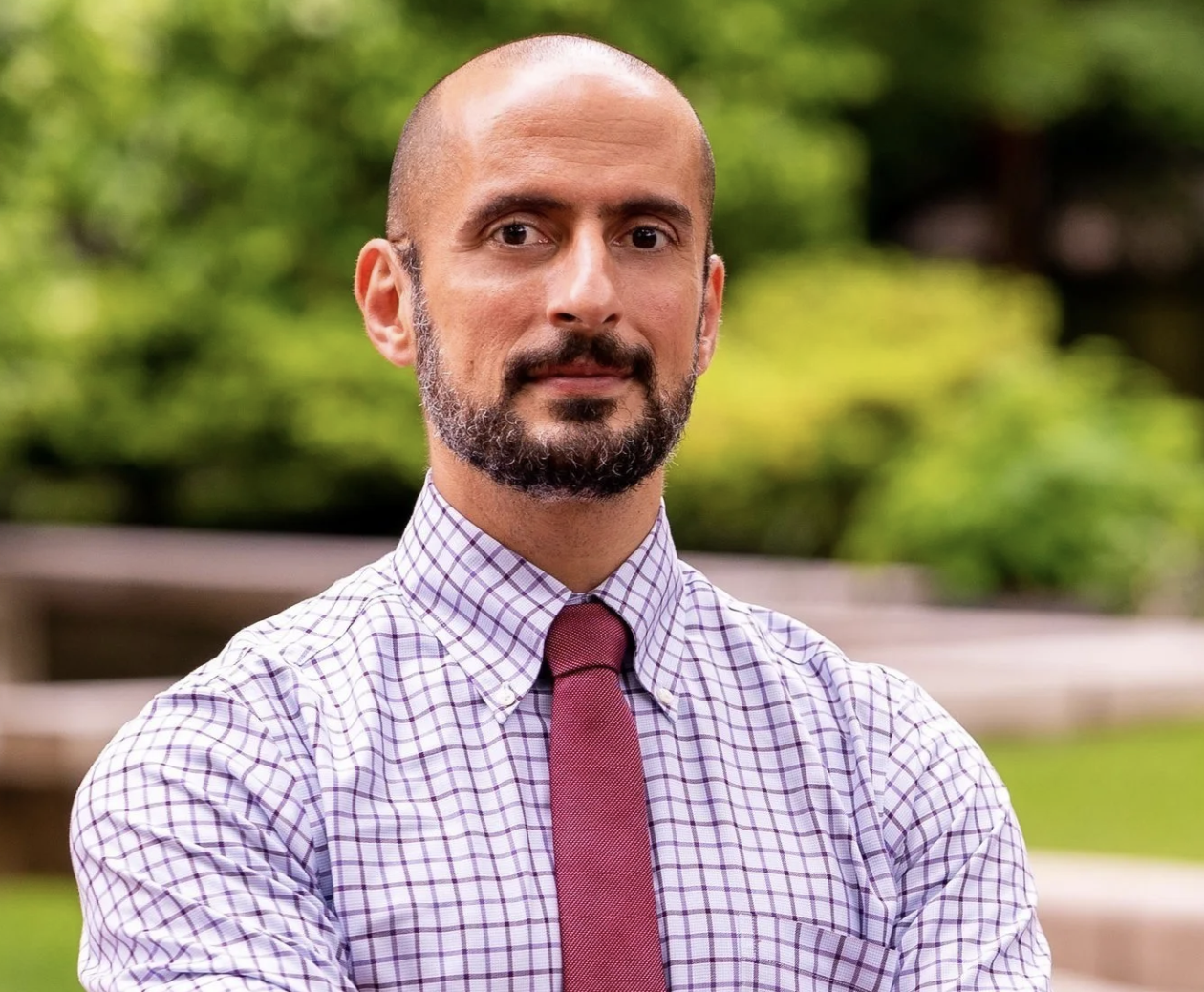 A man with a beard and mustache, wearing a checkered shirt and a maroon tie, standing outdoors with blurred green foliage in the background.