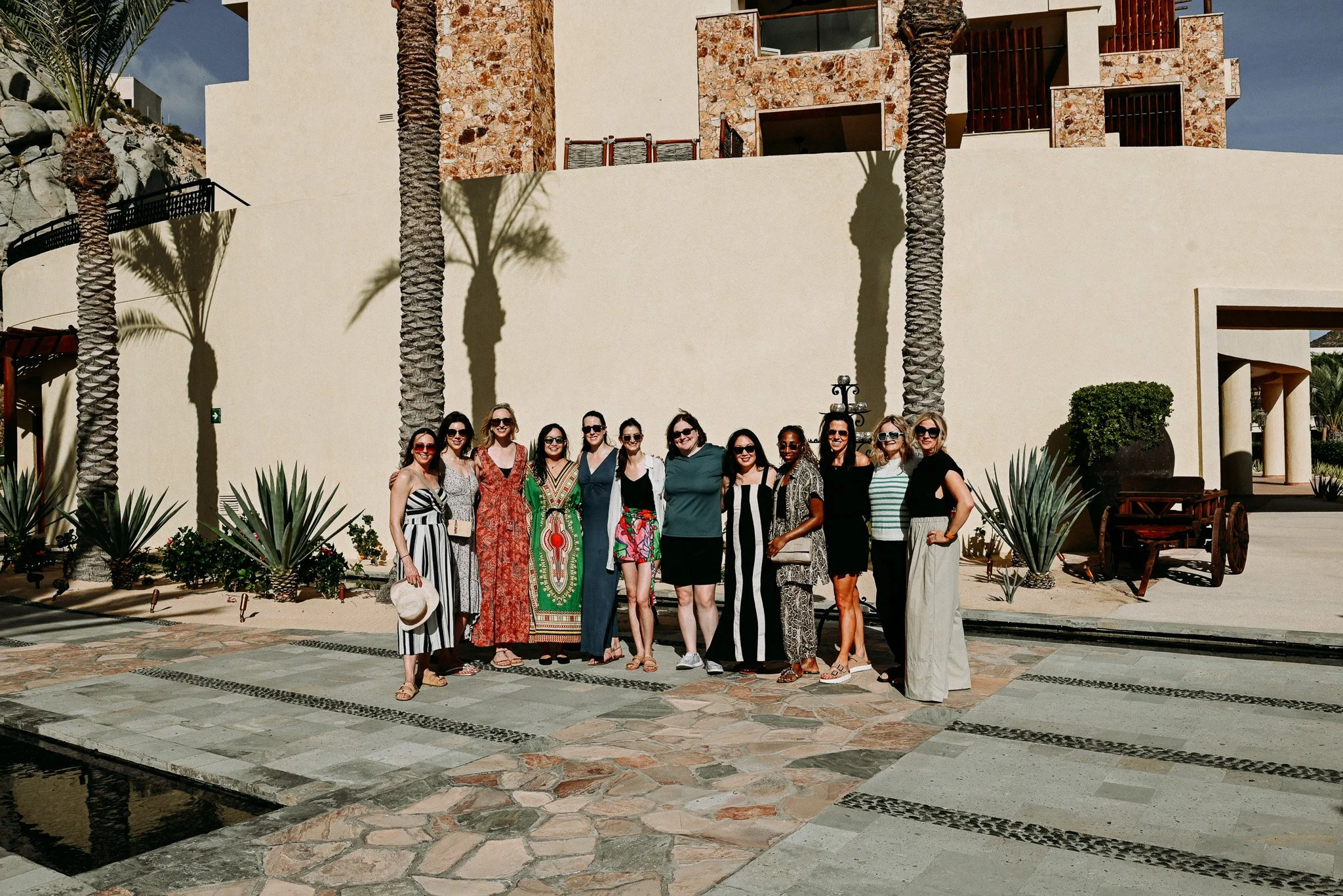 A group of women standing together outdoors in front of a modern building with palm trees, posing for a photo on a sunny day.