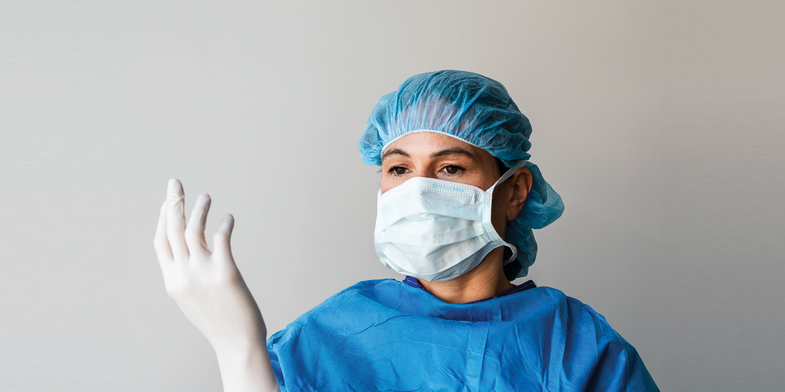 A female healthcare professional wearing a blue surgical cap, a white face mask, and a blue gown adjusting her glove.