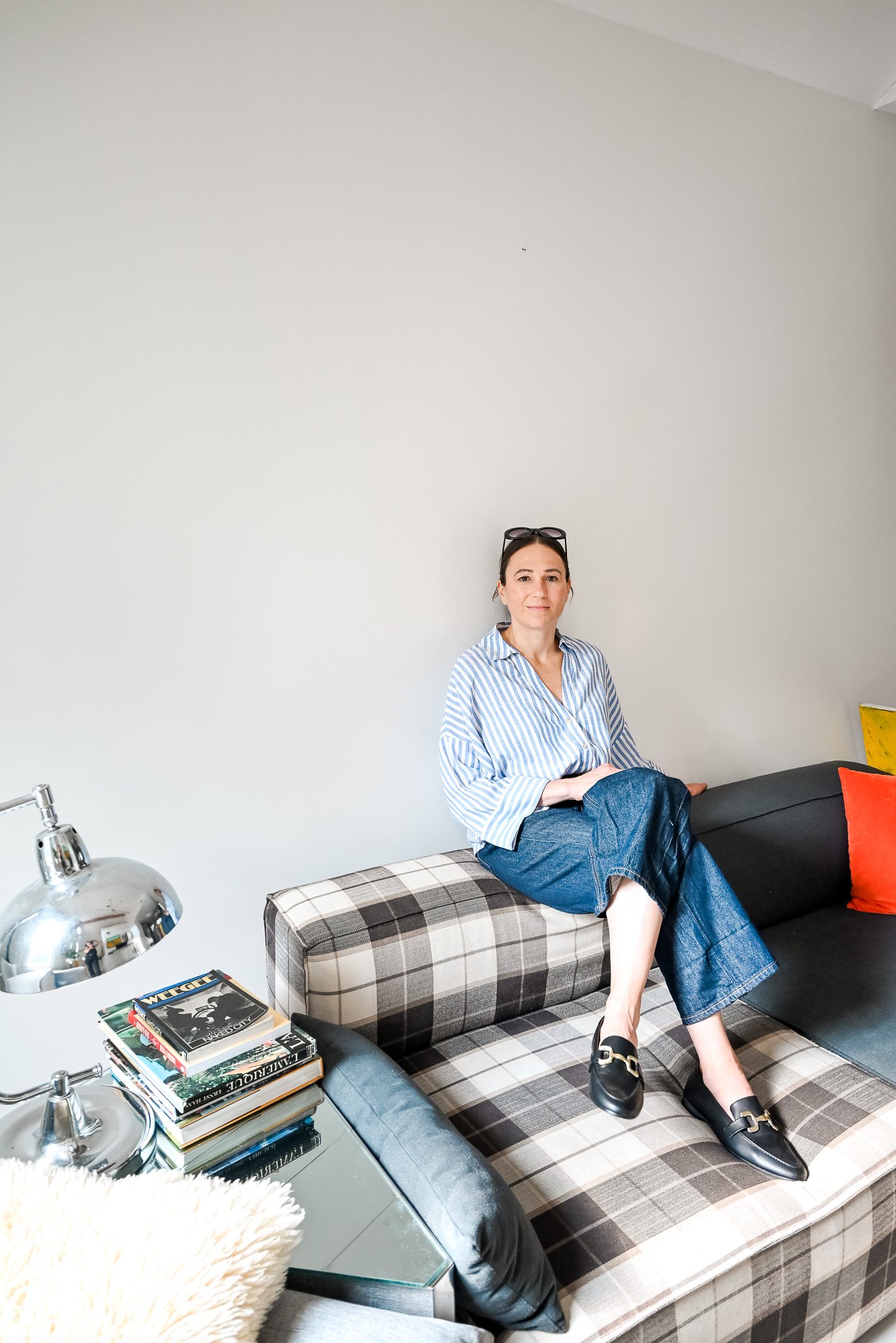 A woman sitting on a plaid-patterned couch with a relaxed pose, wearing a striped shirt, blue jeans, and black loafers, in a room with a minimalist white wall.