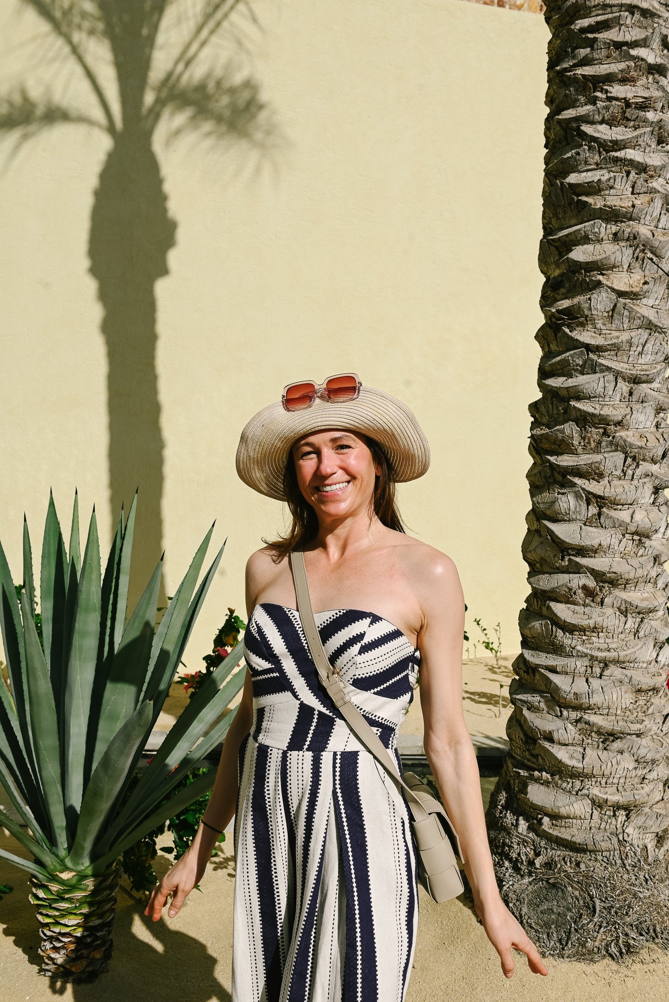 A woman standing outdoors next to a tall palm tree, wearing a striped strapless jumpsuit and a wide-brimmed hat with sunglasses on top, smiling at the camera.