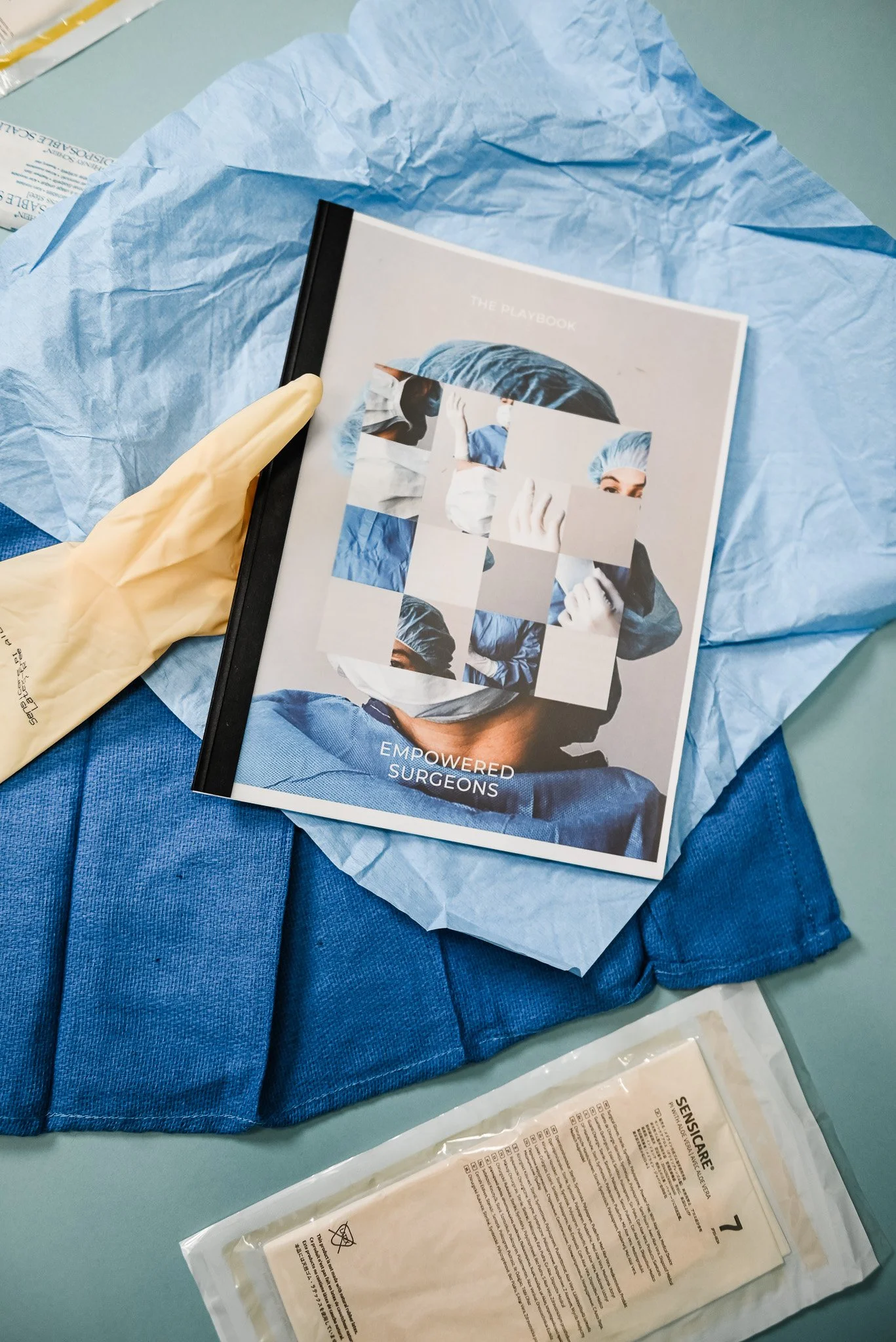 A magazine titled 'The Playbook' with the subtitle 'Empowered Surgeons,' resting on blue surgical drapes. A disposable glove is on the magazine and a sterile medical package is nearby.