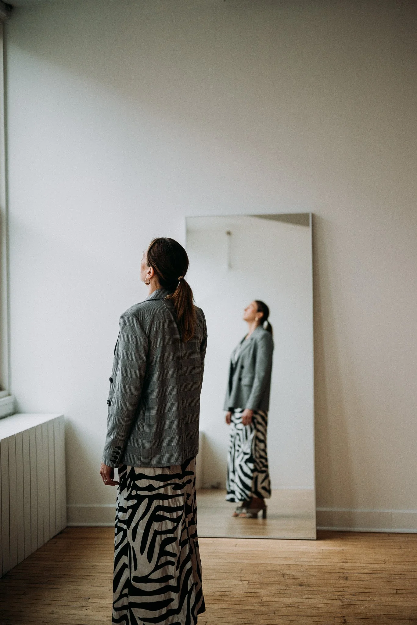 A woman in a grey plaid blazer and zebra print pants stands in front of a mirror, looking to her left in a room with wooden floors and a windowsill.