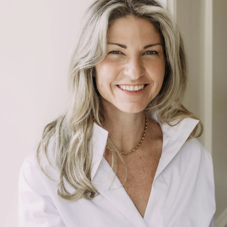 A woman smiling, wearing a white shirt and a gold necklace, with blonde wavy hair, standing in front of a neutral background.