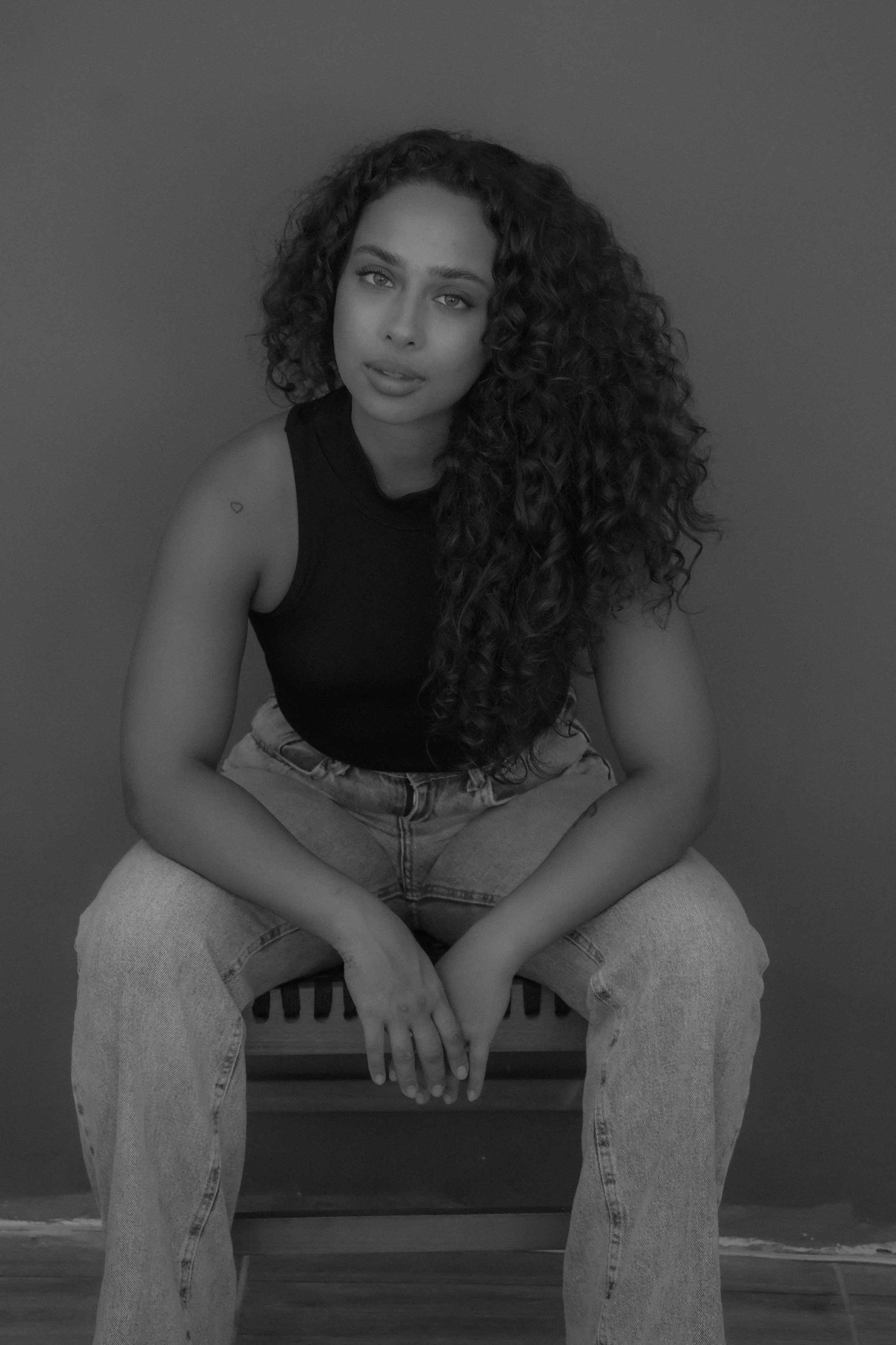A young woman with curly hair and light eyes sits on a wooden chair against a plain wall, wearing a sleeveless top and jeans.