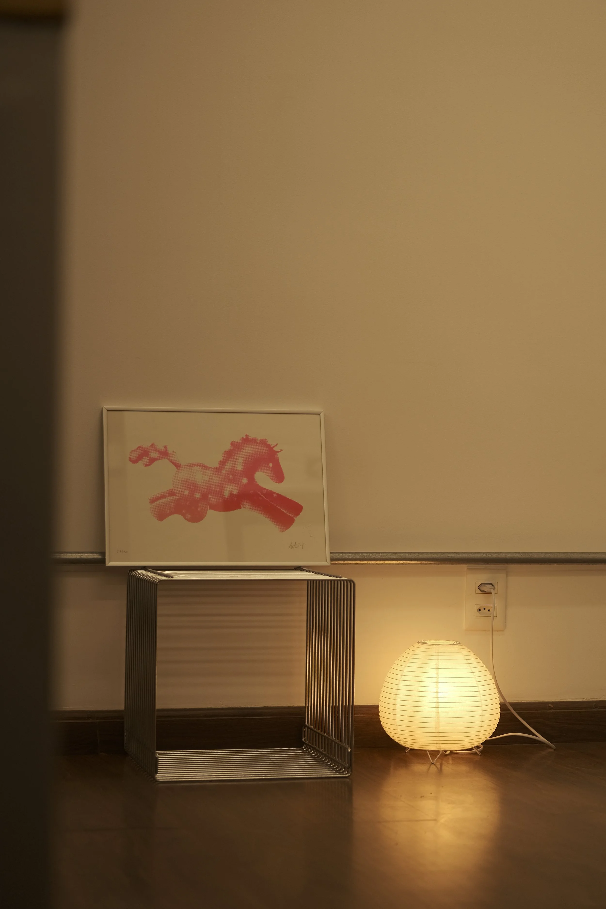 A framed picture of a pink horse with a saddle, a small metal side table, a paper lantern lamp on the floor, and a wall outlet in a cozy room.