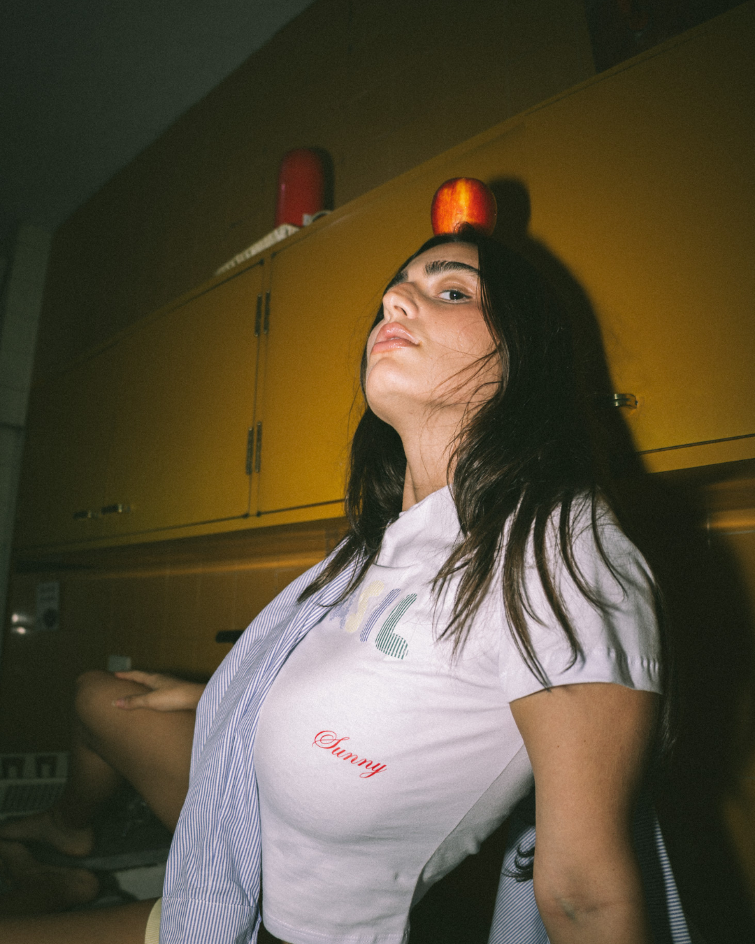 A young woman with dark hair and a serious expression, sitting against a yellow cabinet, with a red apple on her head.