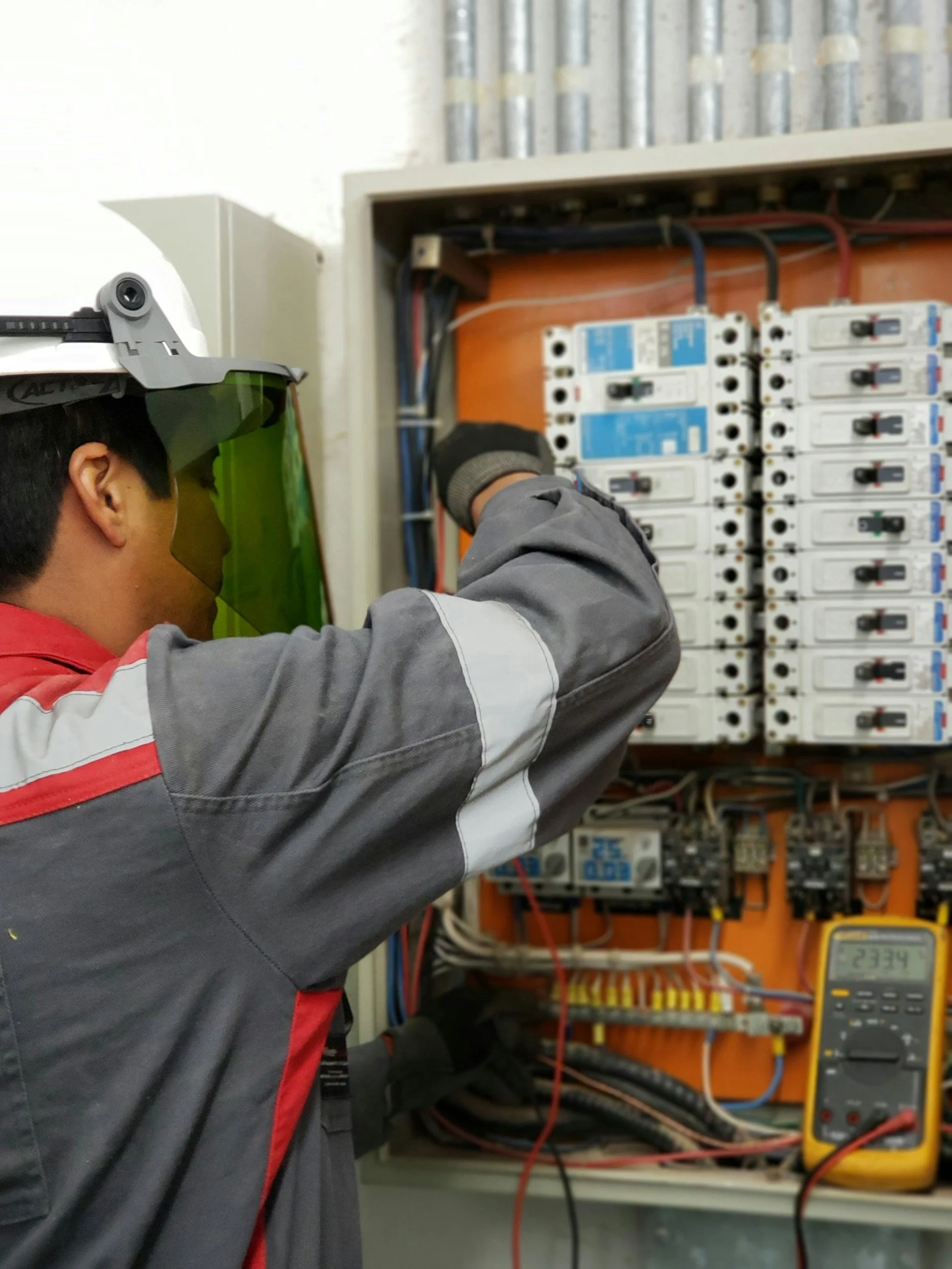 An electrician/ electrical engineer working on an electrical panel, wearing safety goggles and gloves, using a multimeter to test voltage or continuity.