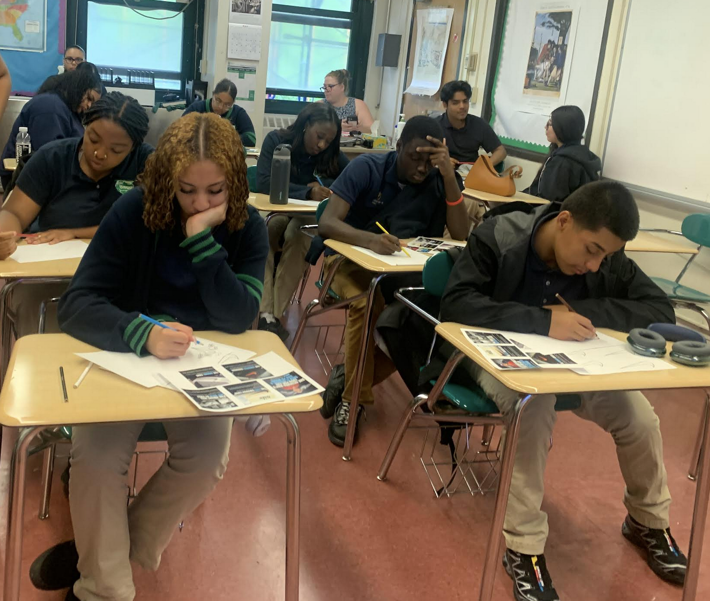 Students sitting at desks in a classroom, focused on writing or drawing on paper, with magazines and stationery on their desks.