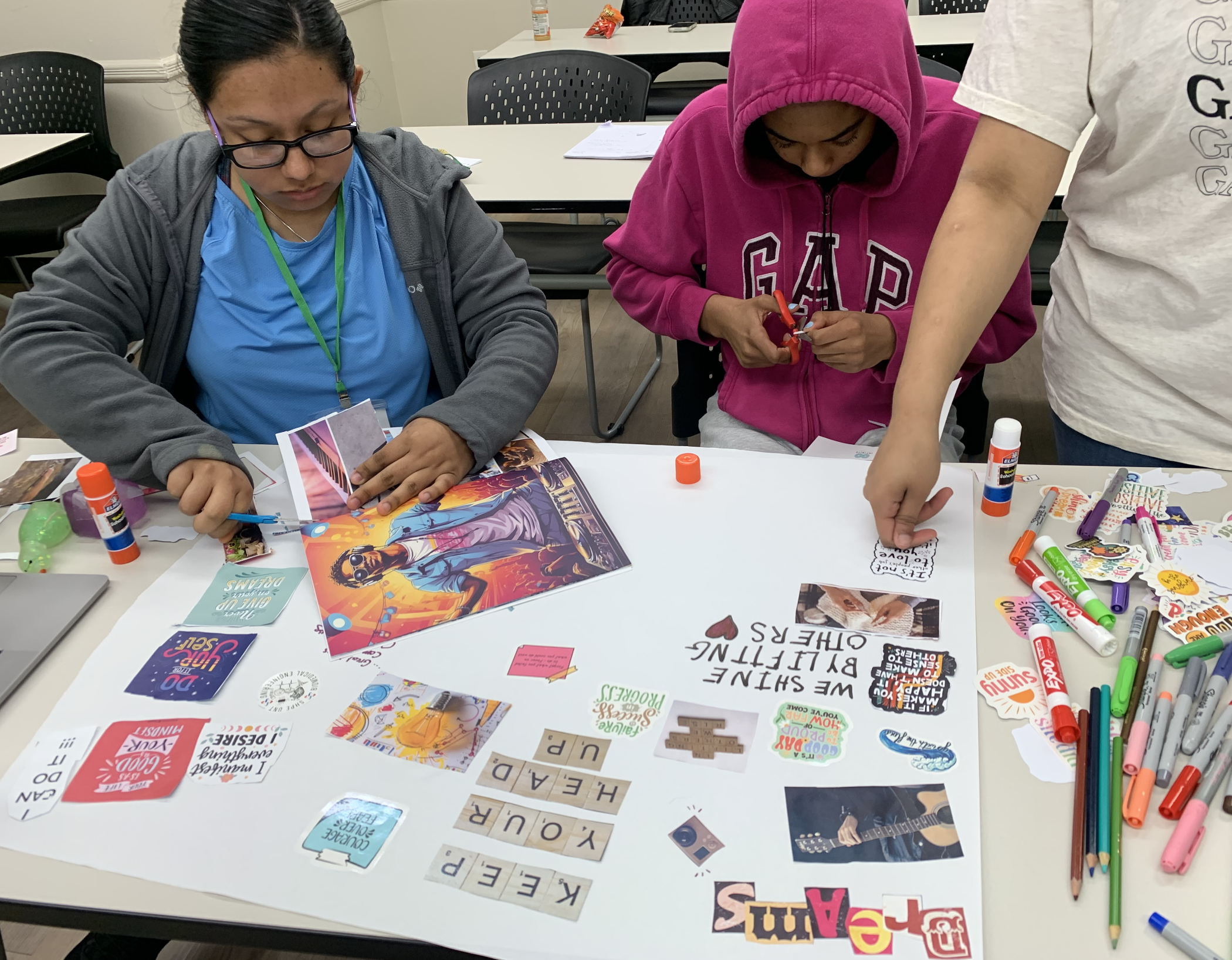 Group of three people working on an arts and crafts project at a table, surrounded by glue sticks, markers, cut-out words, and images.