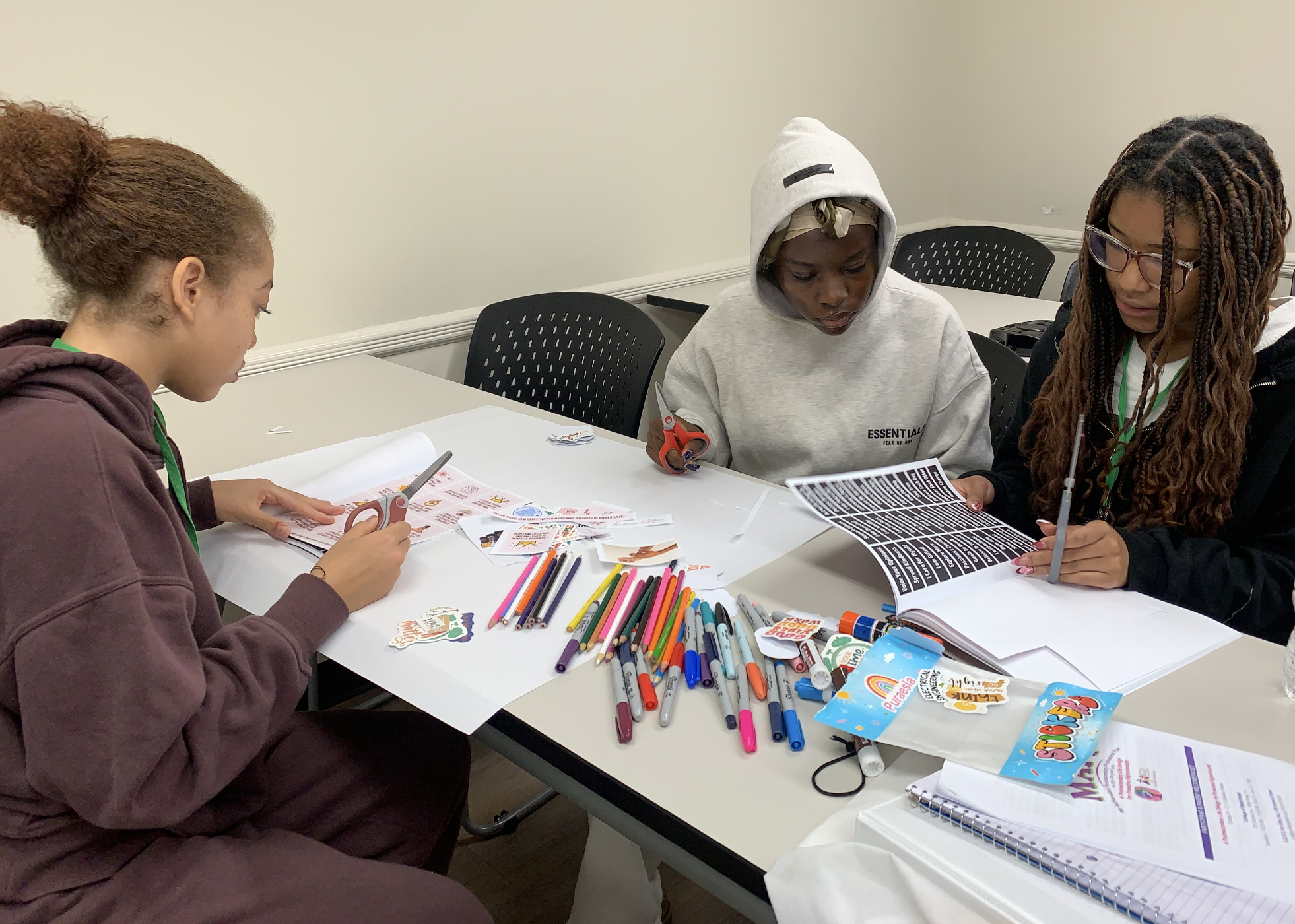 Three young women are seated at a white table, engaged in arts and crafts. The table is cluttered with colored markers, scissors, stickers, and papers. One woman on the left is wearing a maroon hoodie, the woman in the middle has a gray hoodie with a hood and a headscarf, and the woman on the right has glasses and long braided hair. They are focused on their projects. The background is a plain light-colored wall.