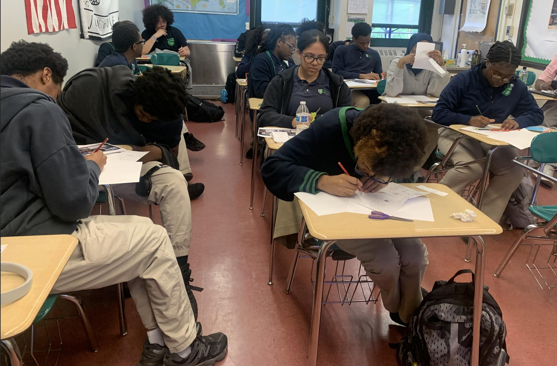 Students in a classroom working on assignments at their desks, some writing, others reading or looking at papers.